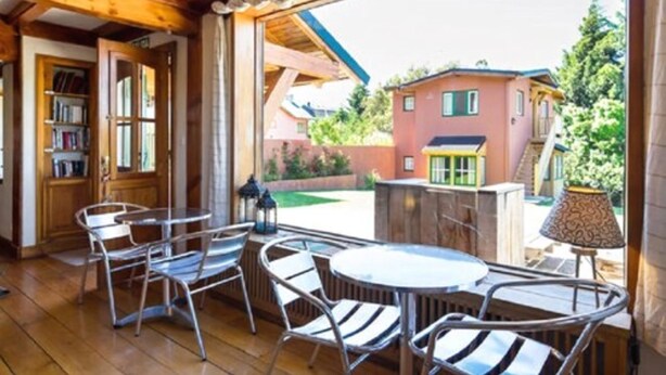 A wooden interior with a patio area featuring metal chairs and a table, a bookshelf, and a view of a red house outside. - La Barraca Suites (San Carlos de Bariloche)
