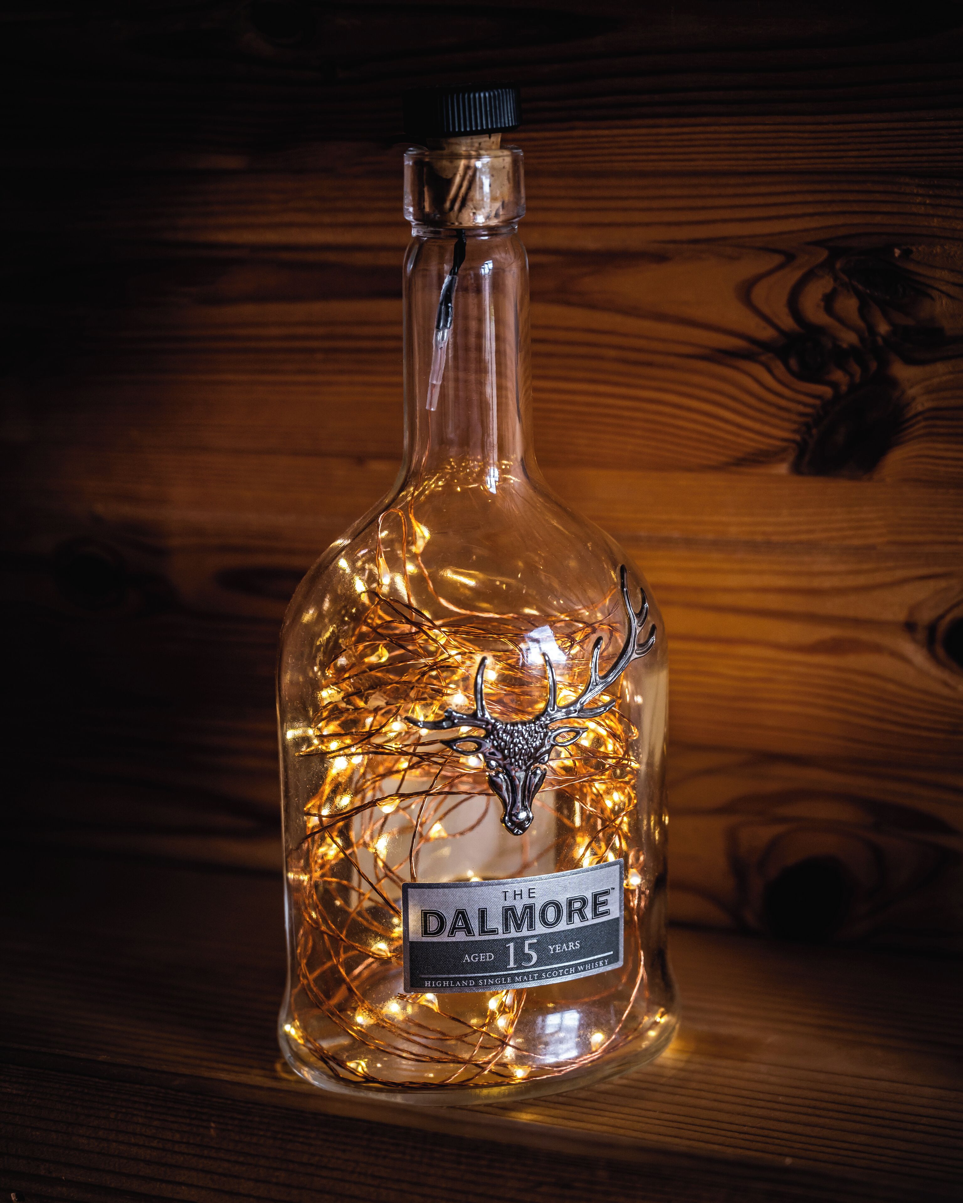 A bottle of Dalmore whisky with a deer head ornament and string lights inside.