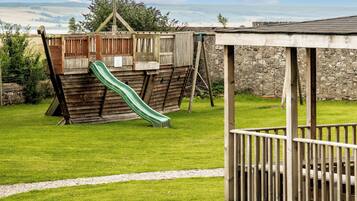 Un parque infantil con tobogán y estructura de escalada, rodeado de césped y un muro de piedra.