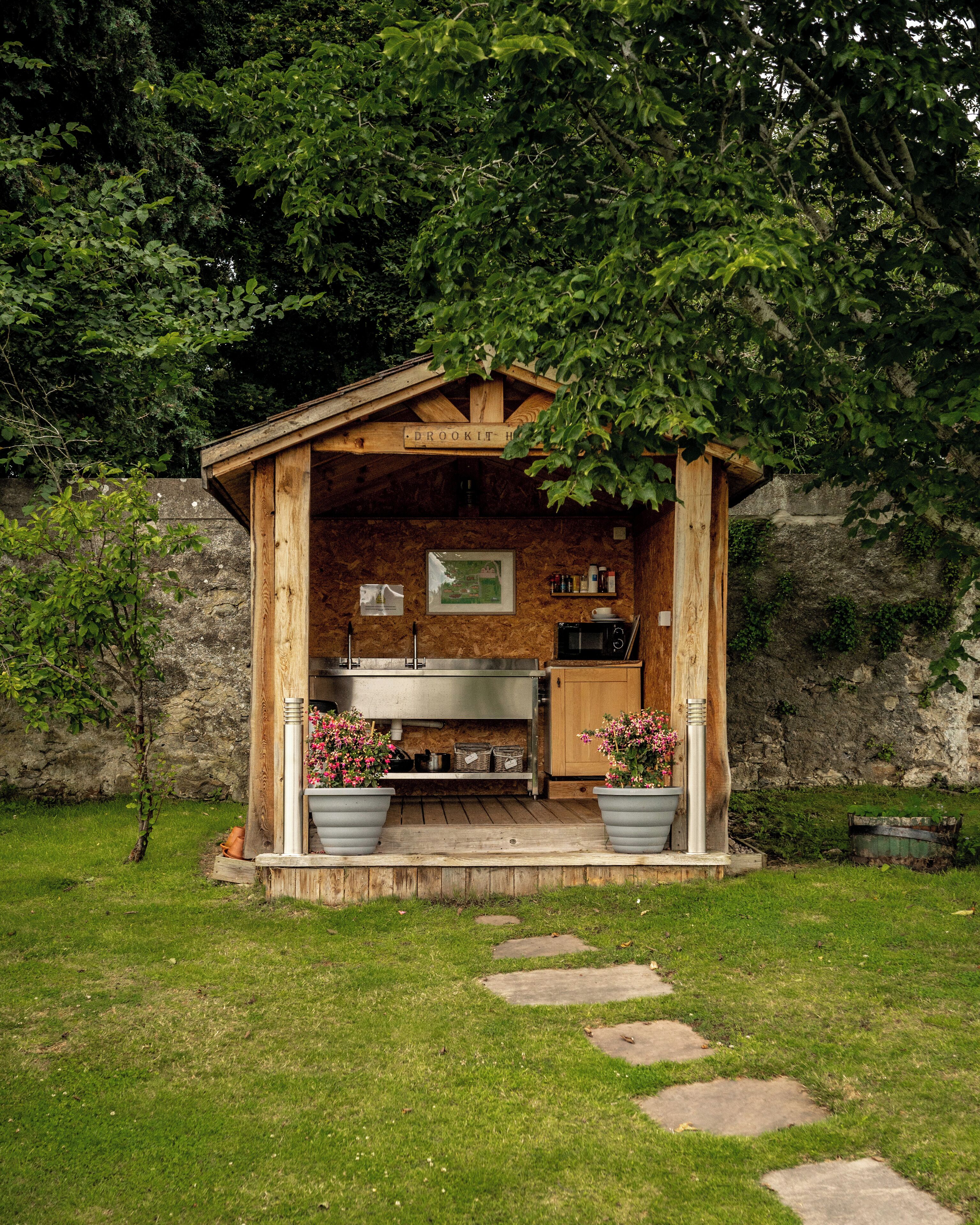 Estructura de madera rústica con zona de cocina, que incluye estufa, pileta y alacenas. Hay plantas en macetas con flores rosas al frente.