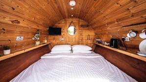 A cozy cabin-style room with a wooden ceiling, a bed with white bedding, a television, a small window, and a shelf with various items. - Delny Glamping (Invergordon)