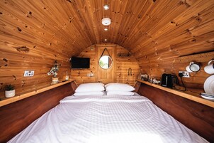 A cozy cabin-style room with a wooden ceiling, a bed with white bedding, a television, a small window, and a shelf with various items.