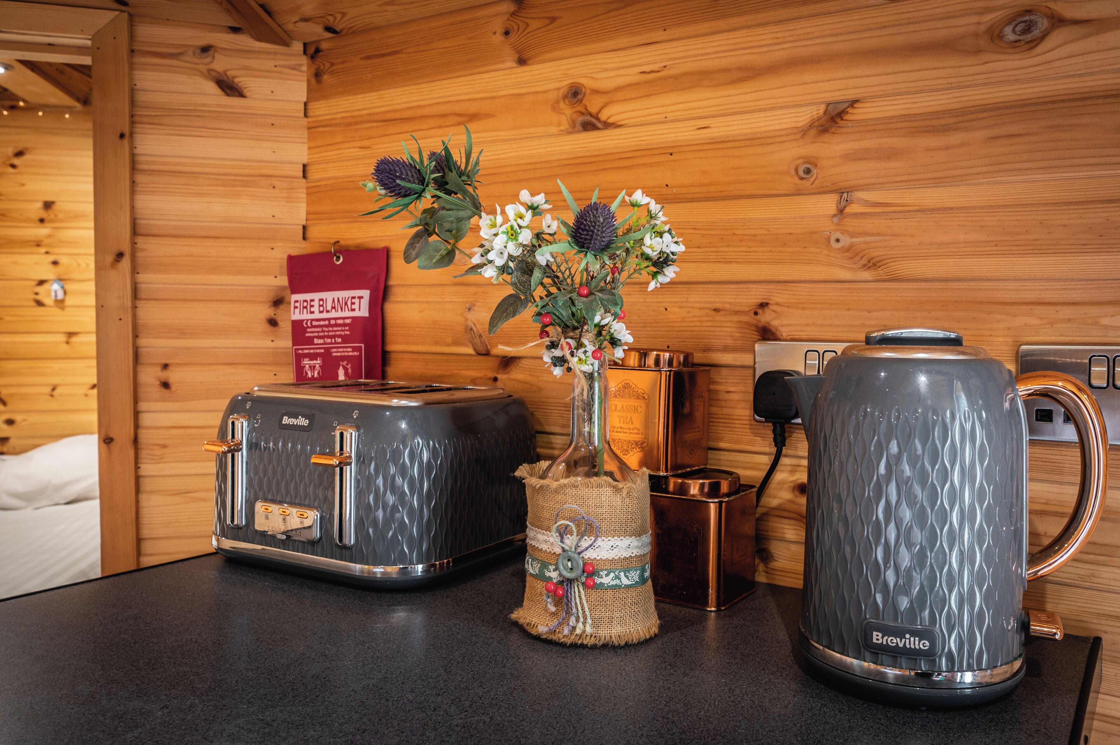 A kitchenette with a Breville toaster, a Breville kettle, and a vase with flowers on a wooden wall.