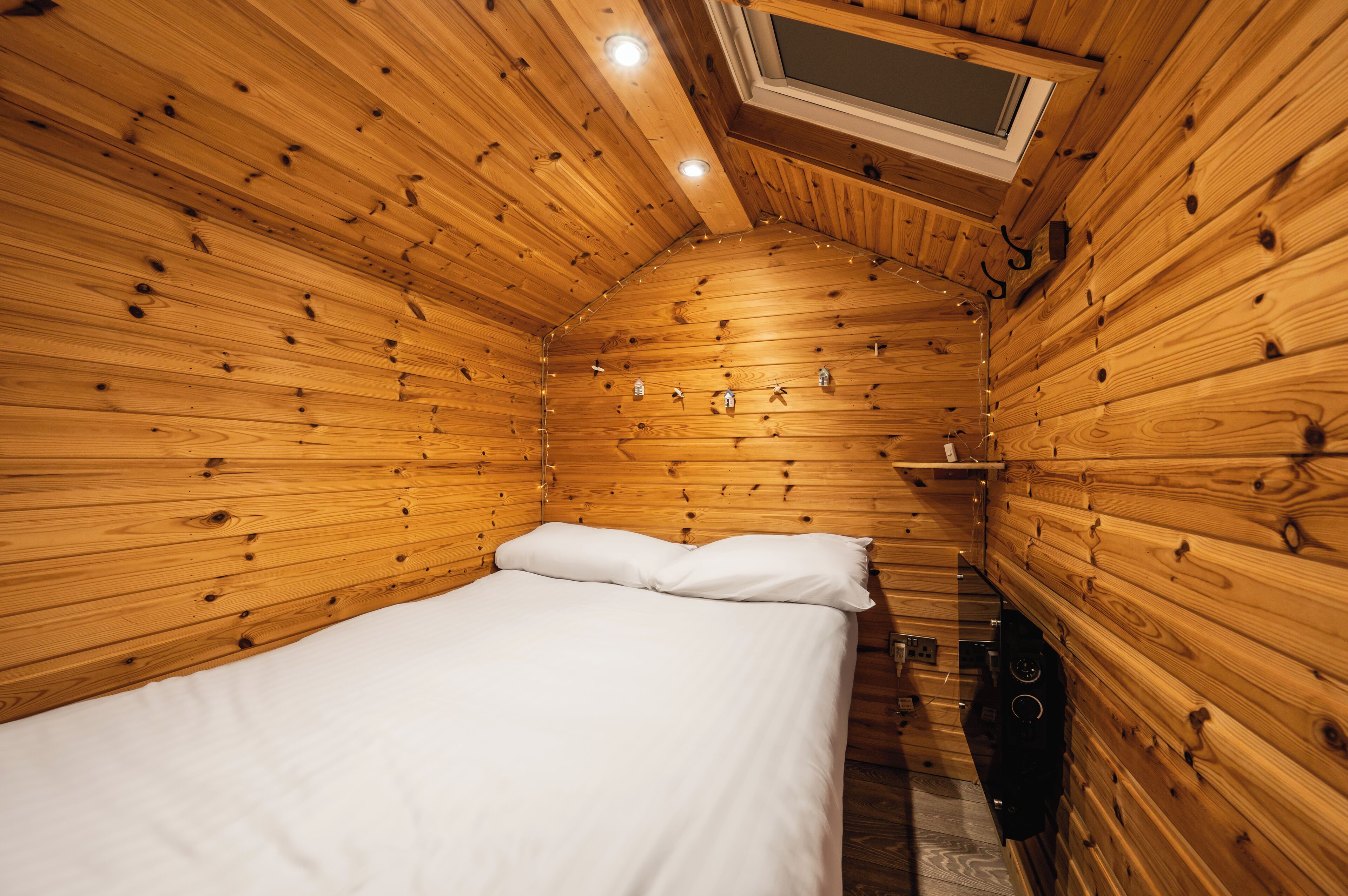 A wooden cabin room with a bed, a small shelf, and a skylight.