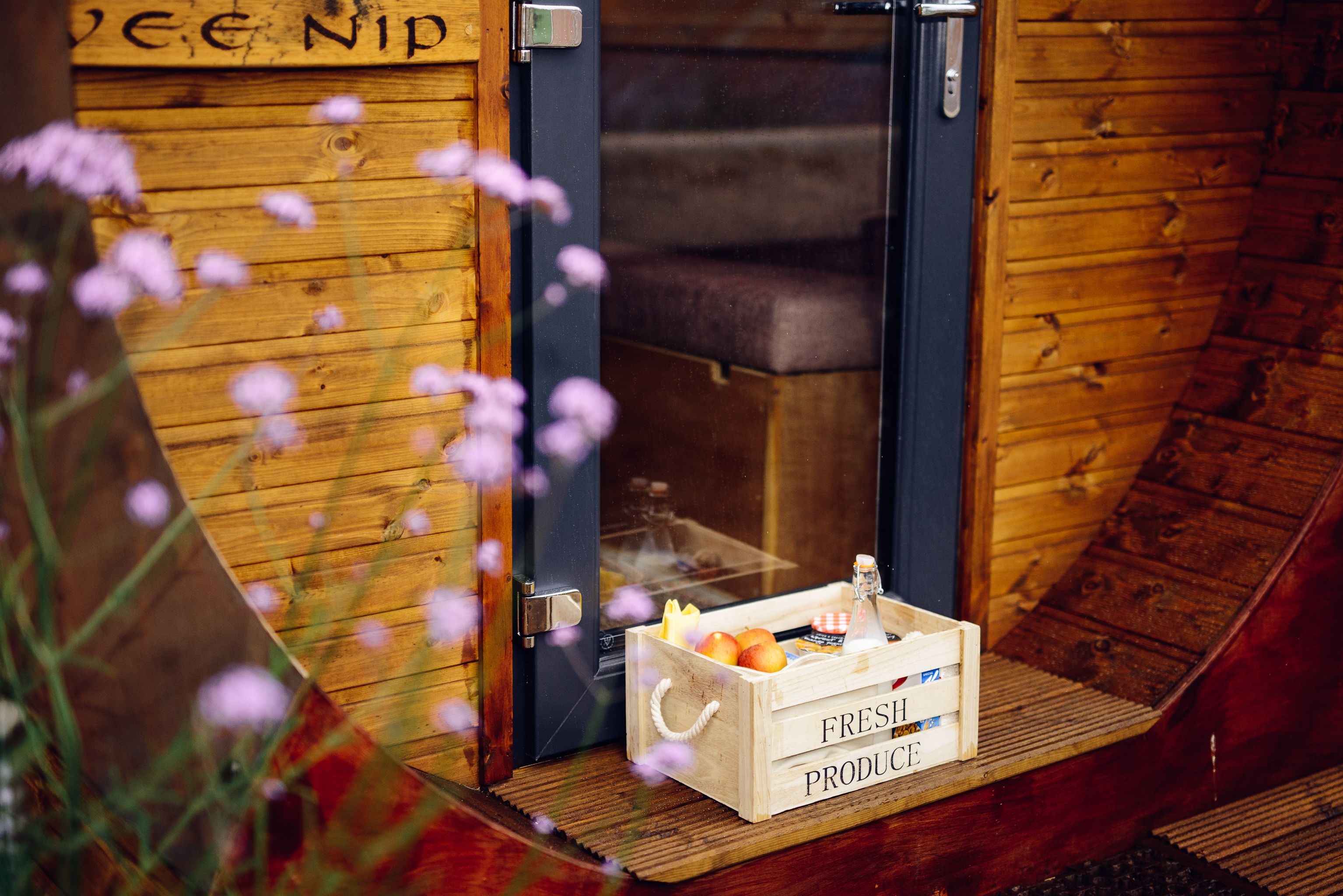 Una porta di legno con un cartello che recita "LEE NIP" e una cassa con l'etichetta "PRODOTTI FRESCHI" contenente frutta e una bottiglia sul portico.