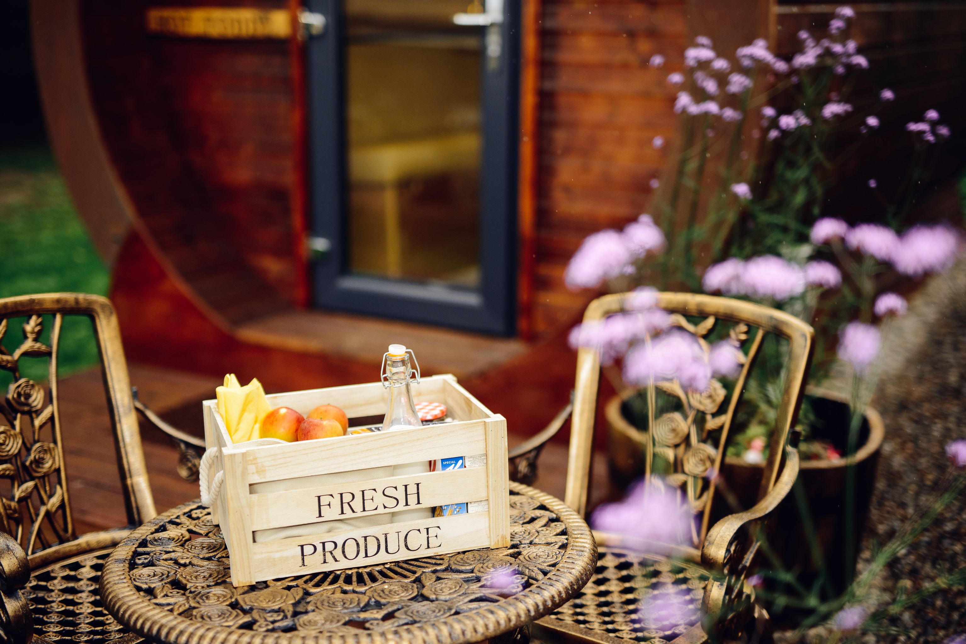 A wooden crate labeled "FRESH PRODUCE" with fruits and a bottle on a round table with ornate chairs.