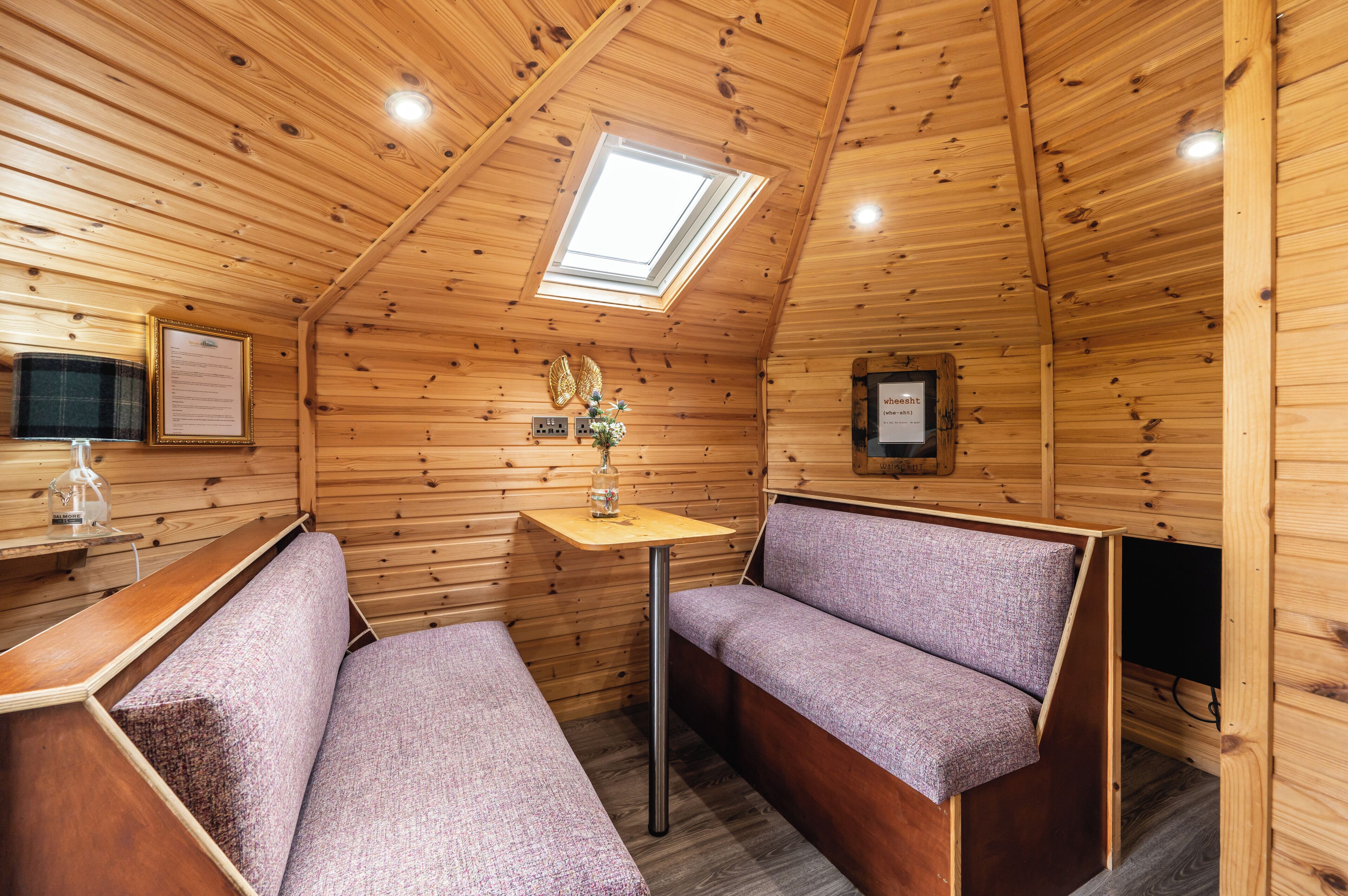 A cozy cabin interior with wooden walls, a skylight, a small table with a vase, and two cushioned seating areas.