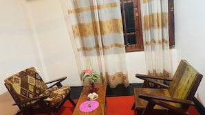 Ein Zimmer mit zwei Holzsesseln, einem kleinen Tisch mit einer pinkfarbenen Matte, einer Topfpflanze und einem Fenster mit Vorhängen.