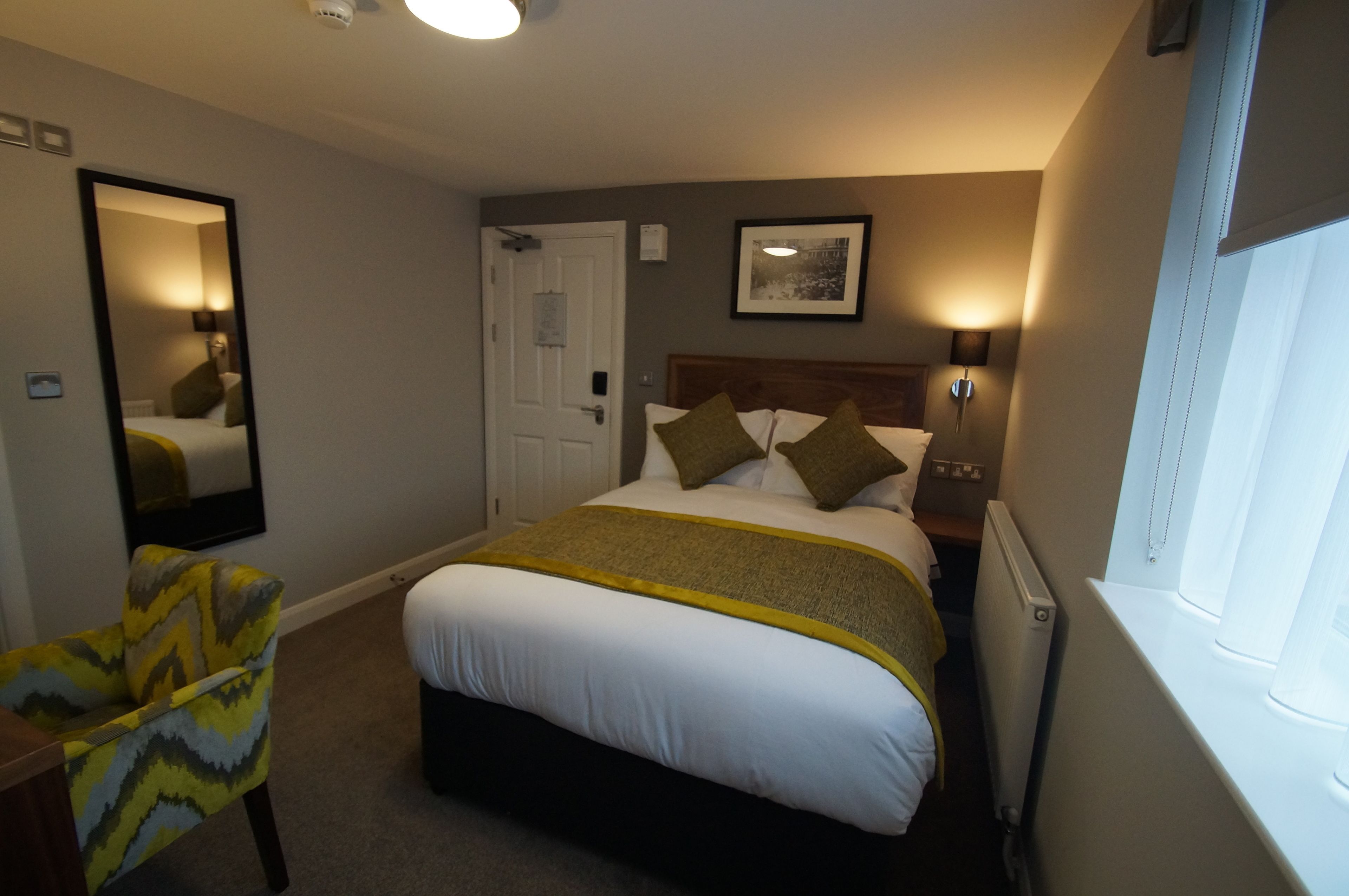 A hotel room with a bed, a chair, a bedside table, a mirror, a window, and a door.