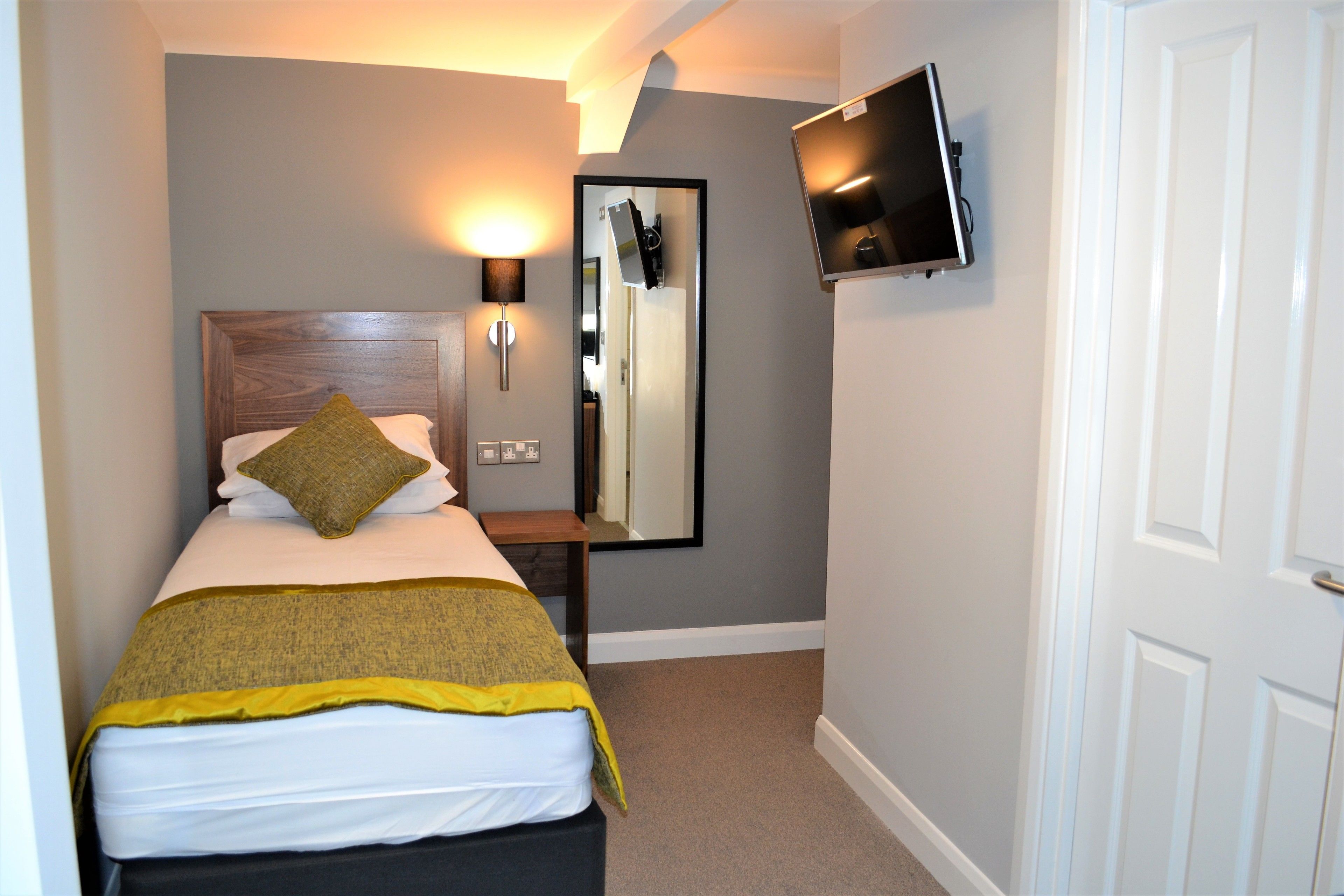 A hotel room with a bed, a bedside table, a mirror, a TV mounted on the wall, and a door.
