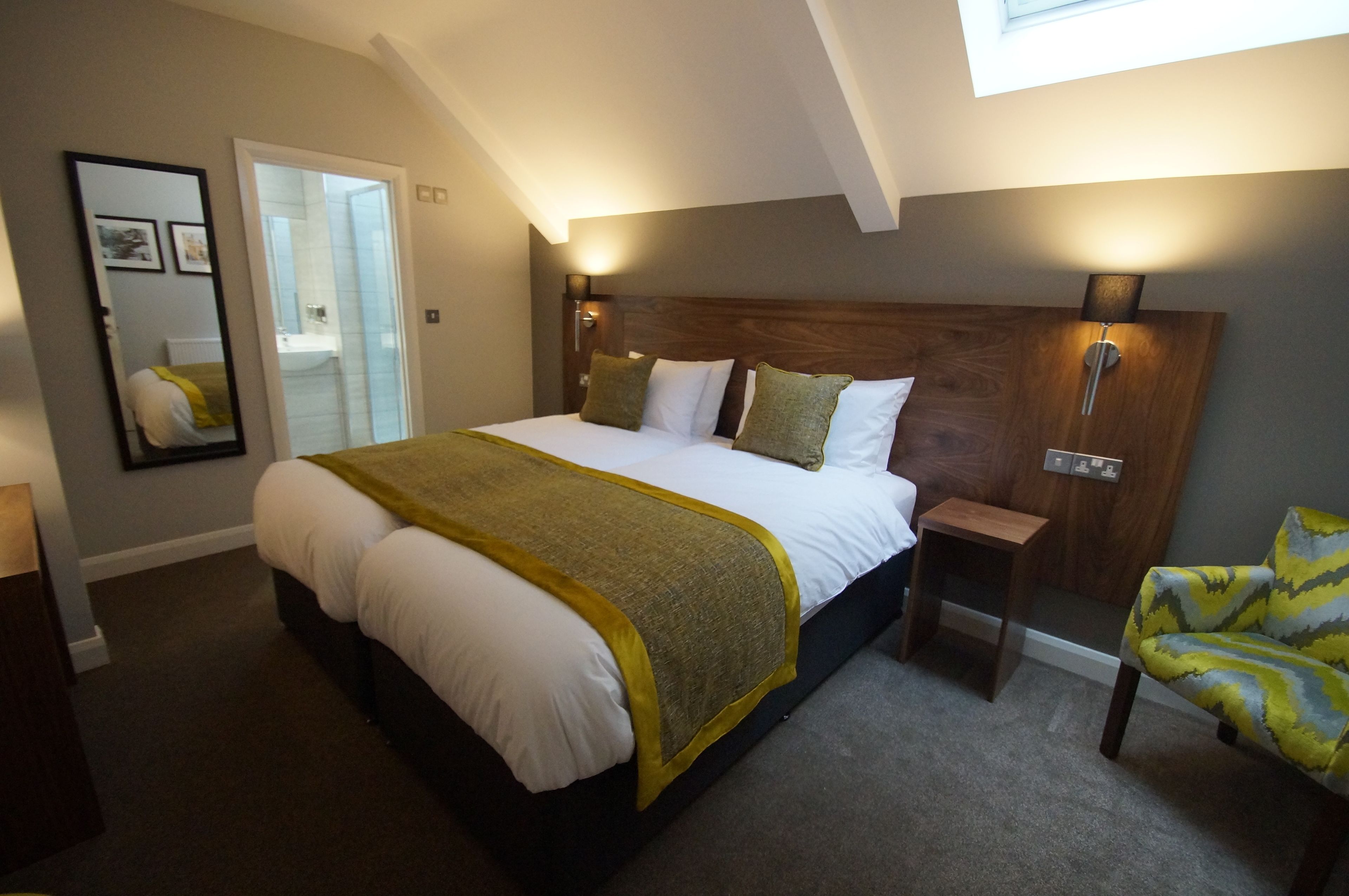 A bedroom with a large bed, a bedside table, a chair, a mirror, and a skylight.