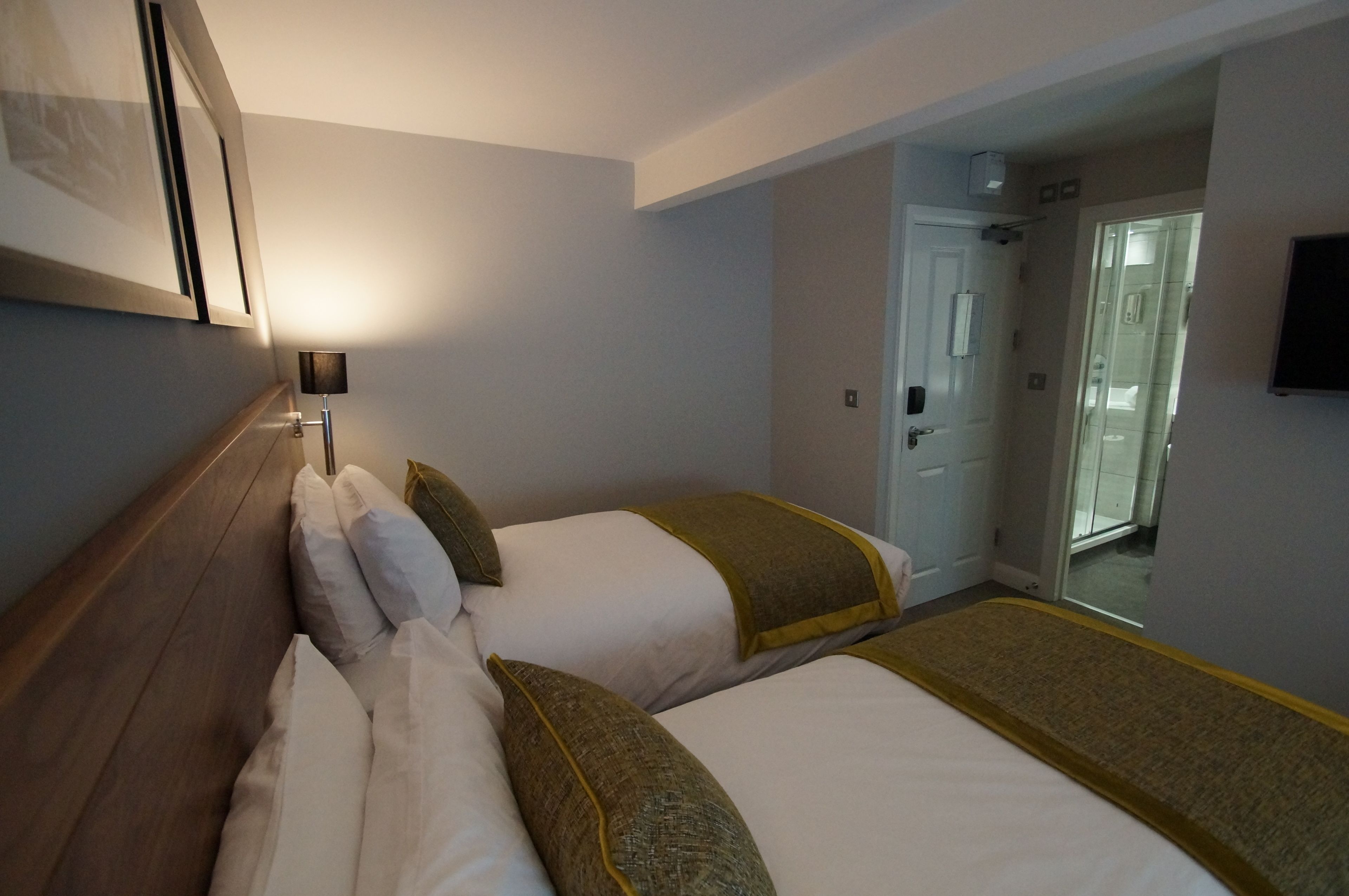 A hotel room with two beds, a bedside table, a lamp, and a television on the wall.
