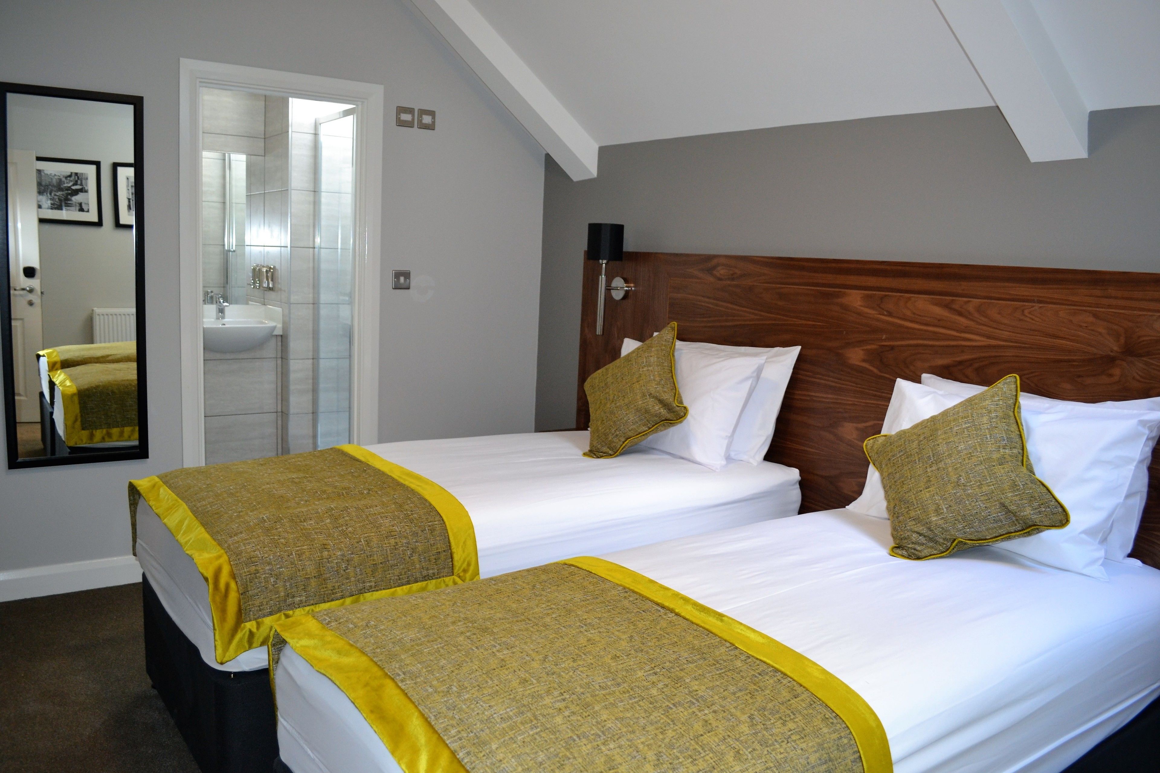 Habitación de hotel con dos camas, cabecera de madera, un espejo y un baño con lavamanos y espejo.