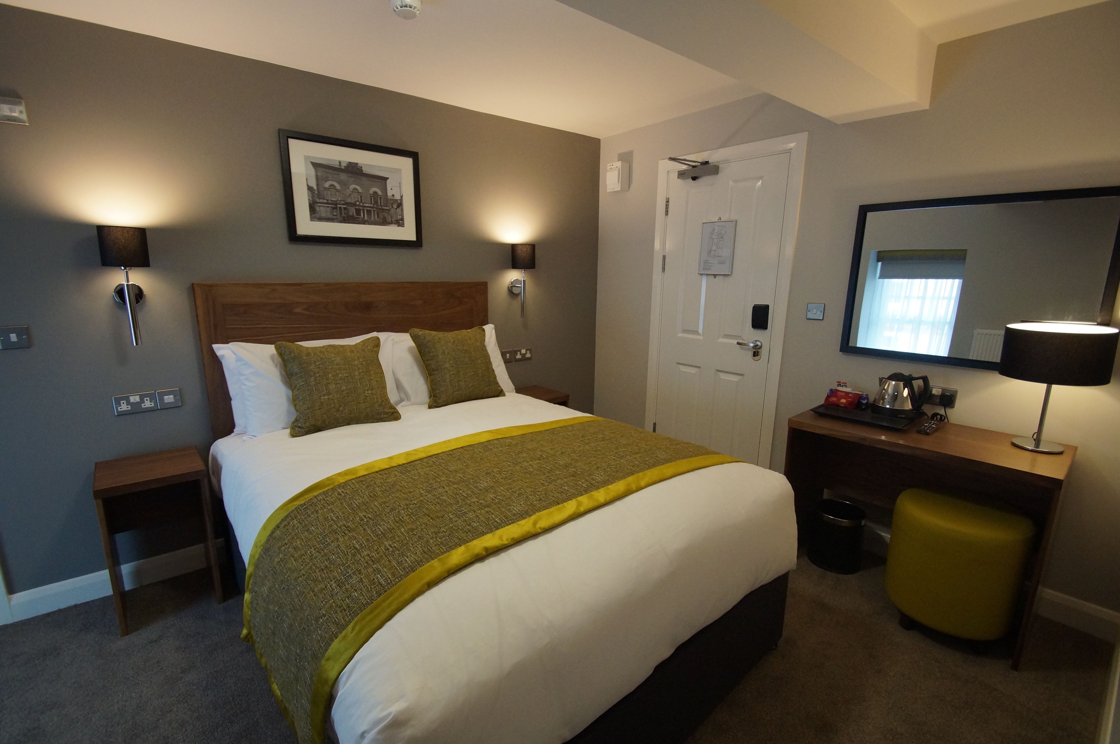 Habitación de hotel ordenada con una cama grande, lámparas de noche, una mesita de noche de madera, un escritorio con silla y un espejo.