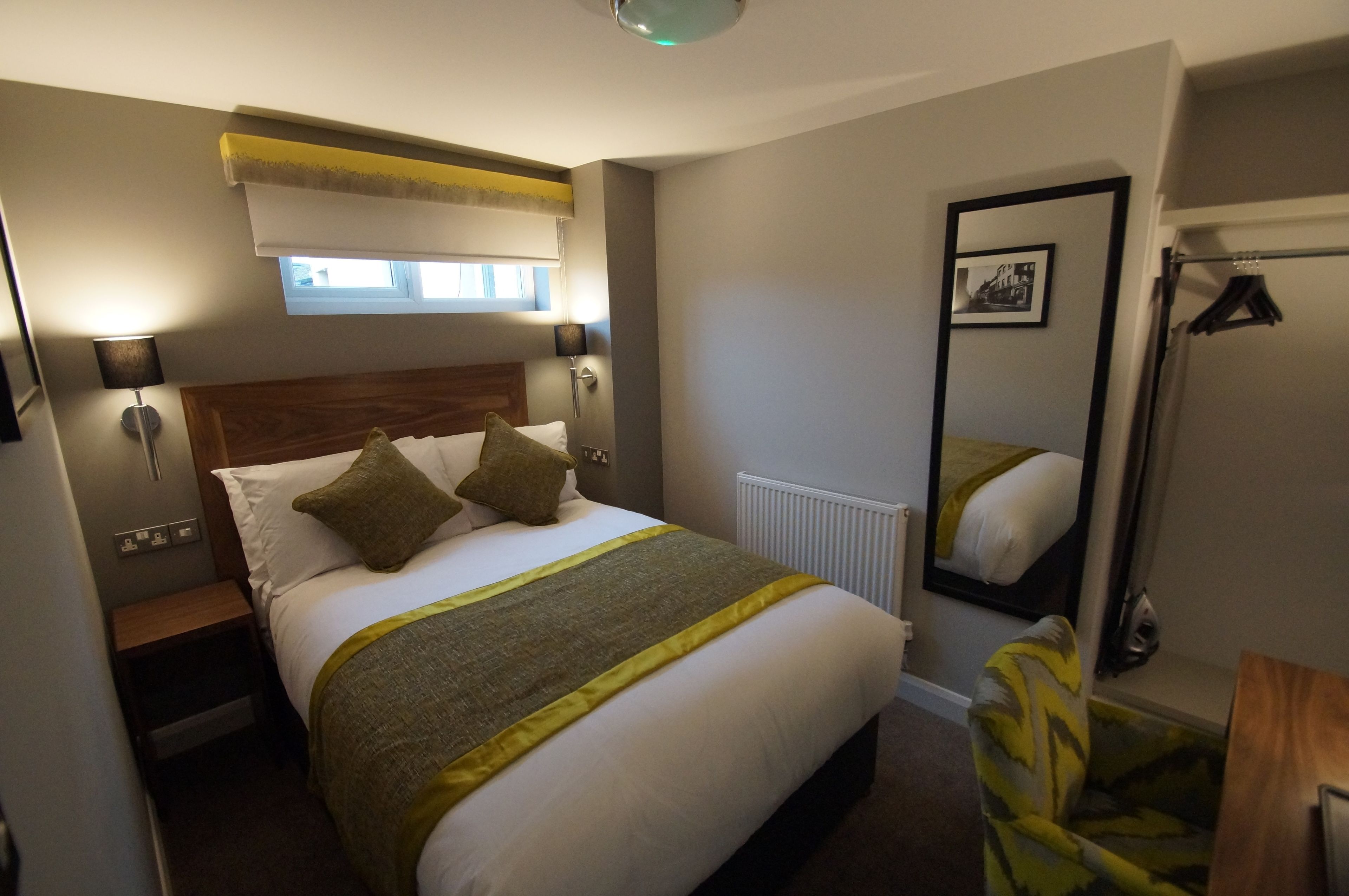 Habitación de hotel ordenada con una cama grande, lámparas de noche, una ventana con persianas, un espejo y un perchero.