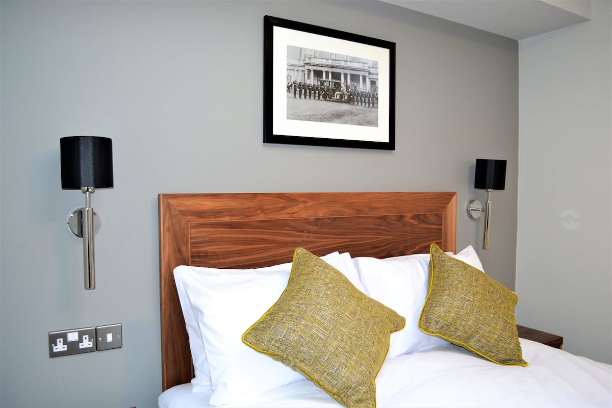 Een hotelkamer met een houten hoofdeinde, twee witte kussens, twee mosterdgele kussens en een ingelijst fotoportret aan de muur.