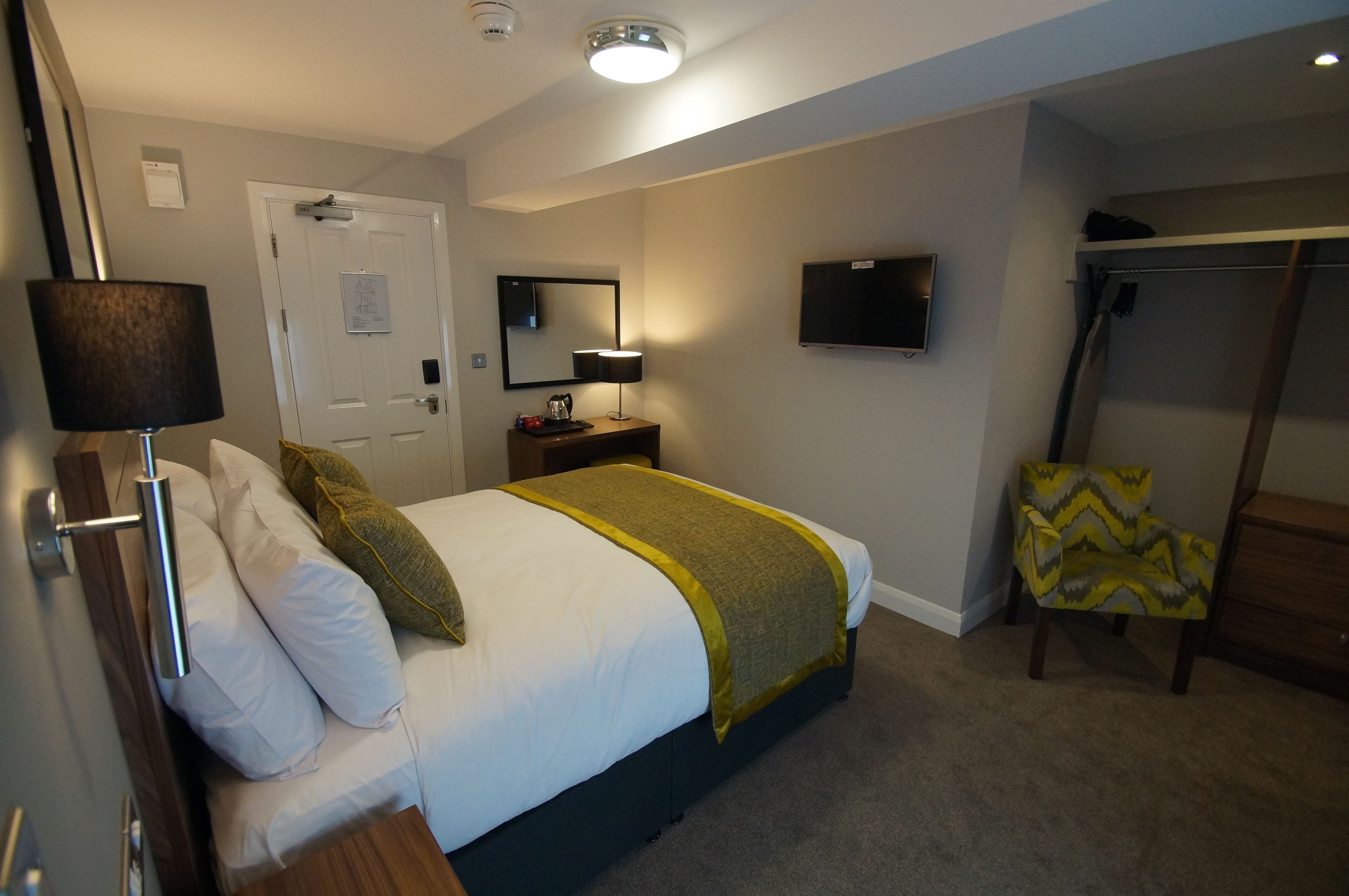 Habitación de hotel con una cama, una mesita de noche, una lámpara, una silla, un televisor y un perchero.