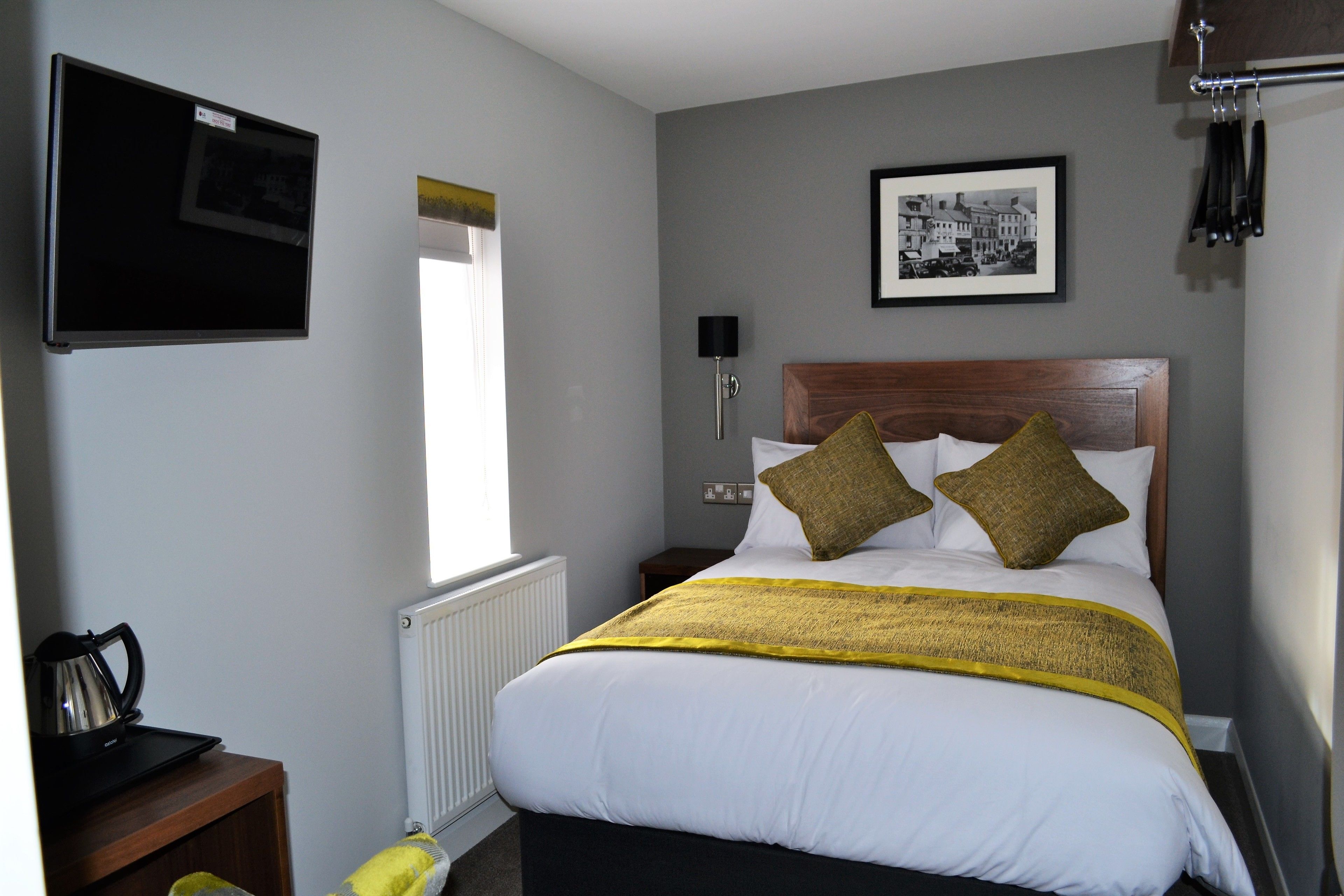 A neatly arranged hotel room with a bed, a bedside table with a kettle, a framed picture on the wall, and a window with a view.