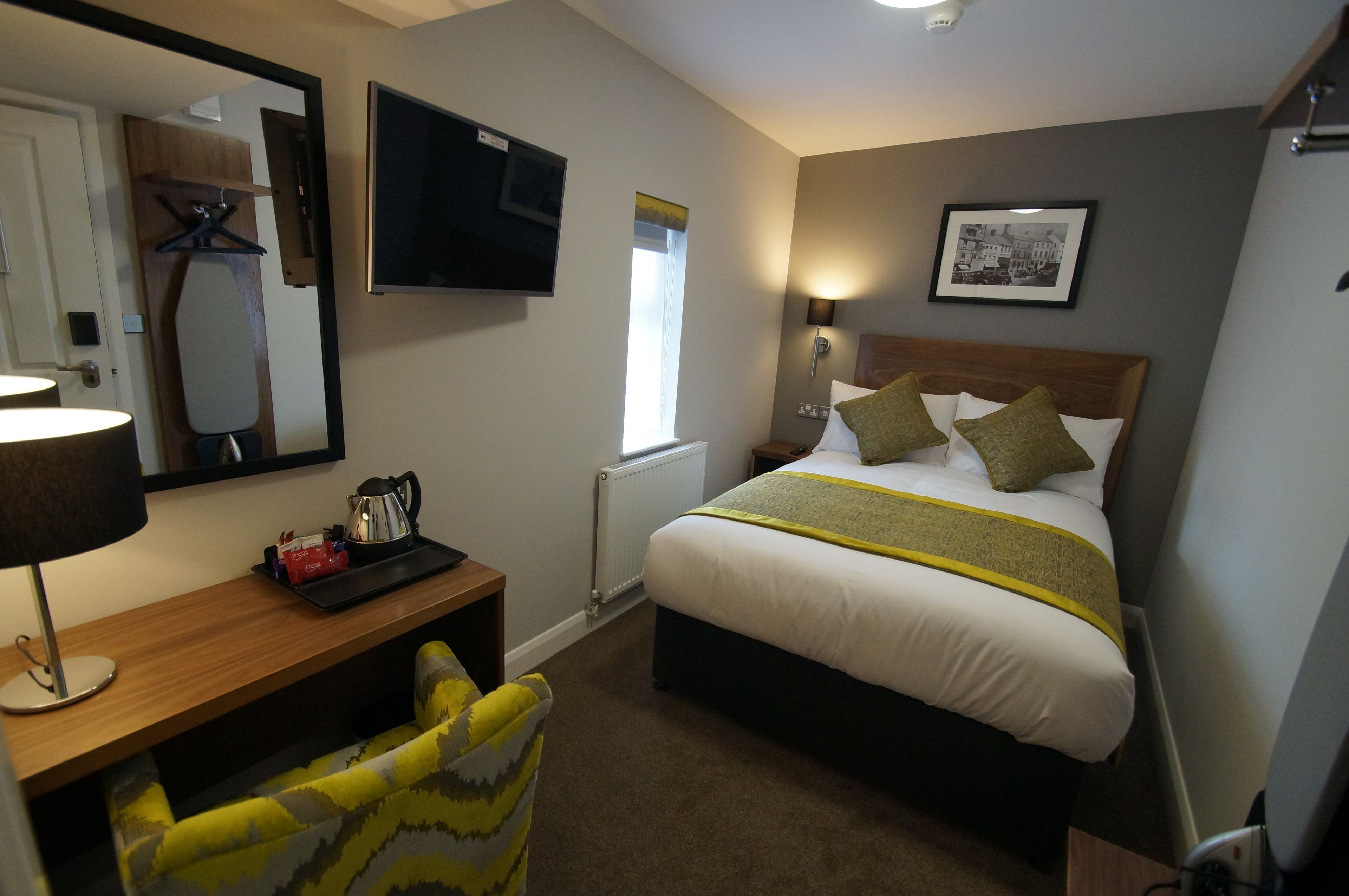 Een hotelkamer met een bed, nachtkastje, lamp, stoel en een televisie aan de muur.