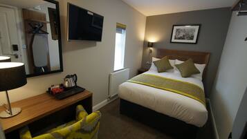 Een hotelkamer met een bed, nachtkastje, lamp, stoel en een televisie aan de muur.