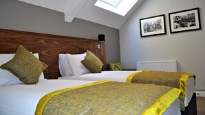 Une chambre d’hôtel avec une tête de lit en bois, deux lits avec une literie jaune et verte, une lucarne et deux photographies en noir et blanc accrochées au mur.