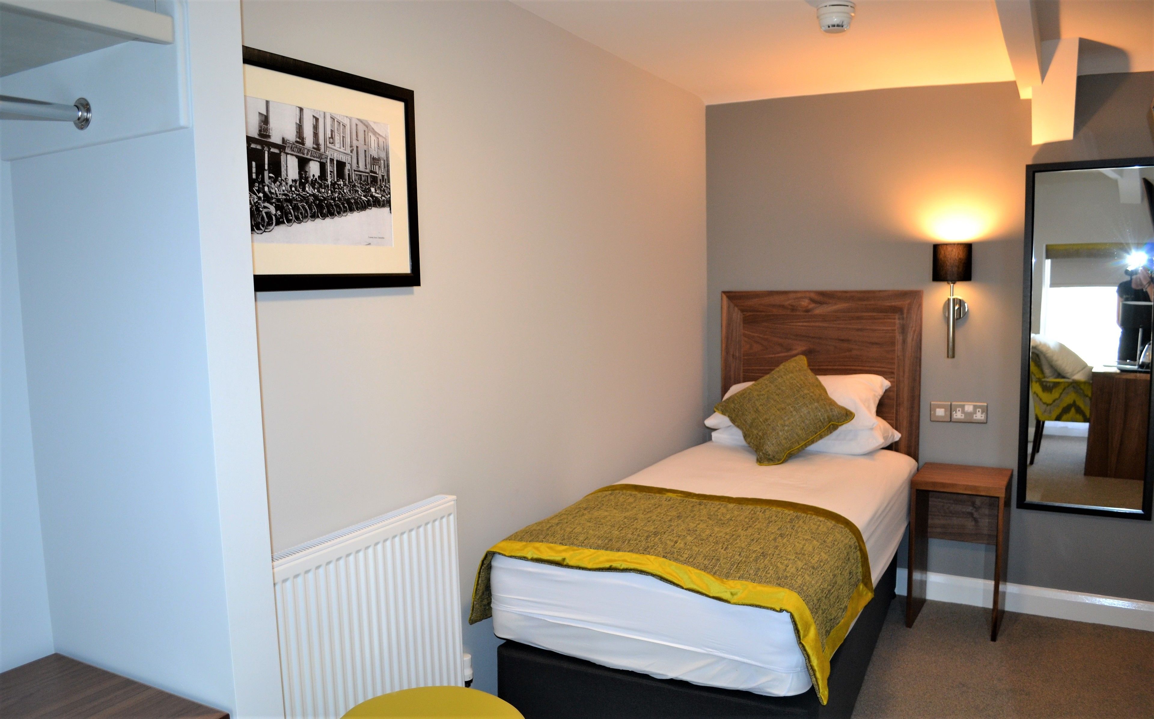 A neatly arranged bedroom with a bed, a bedside table, a lamp, a radiator, and a framed picture on the wall.