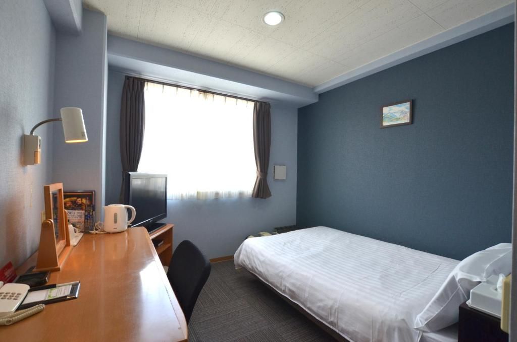 Habitación de hotel con una cama individual, un escritorio con teléfono y cafetera, un televisor y una ventana con cortinas.