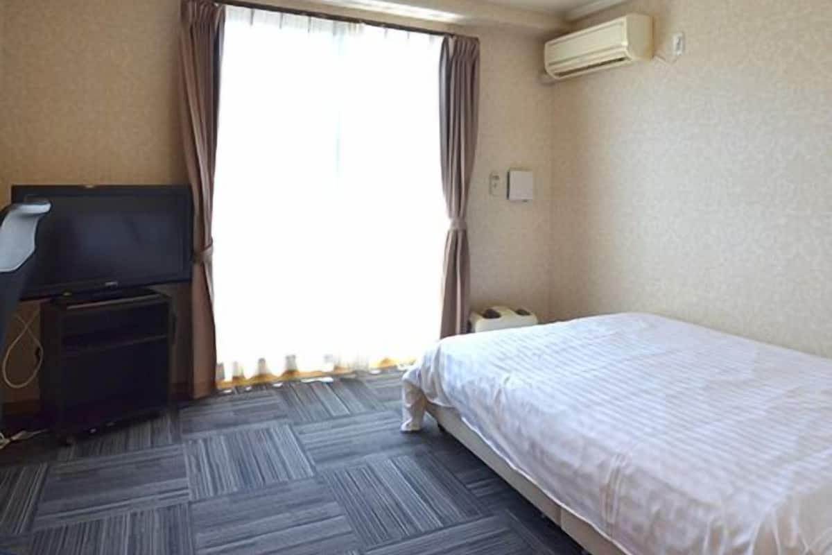 A hotel room with a bed, a desk with a computer, an air conditioning unit, and a window with curtains.