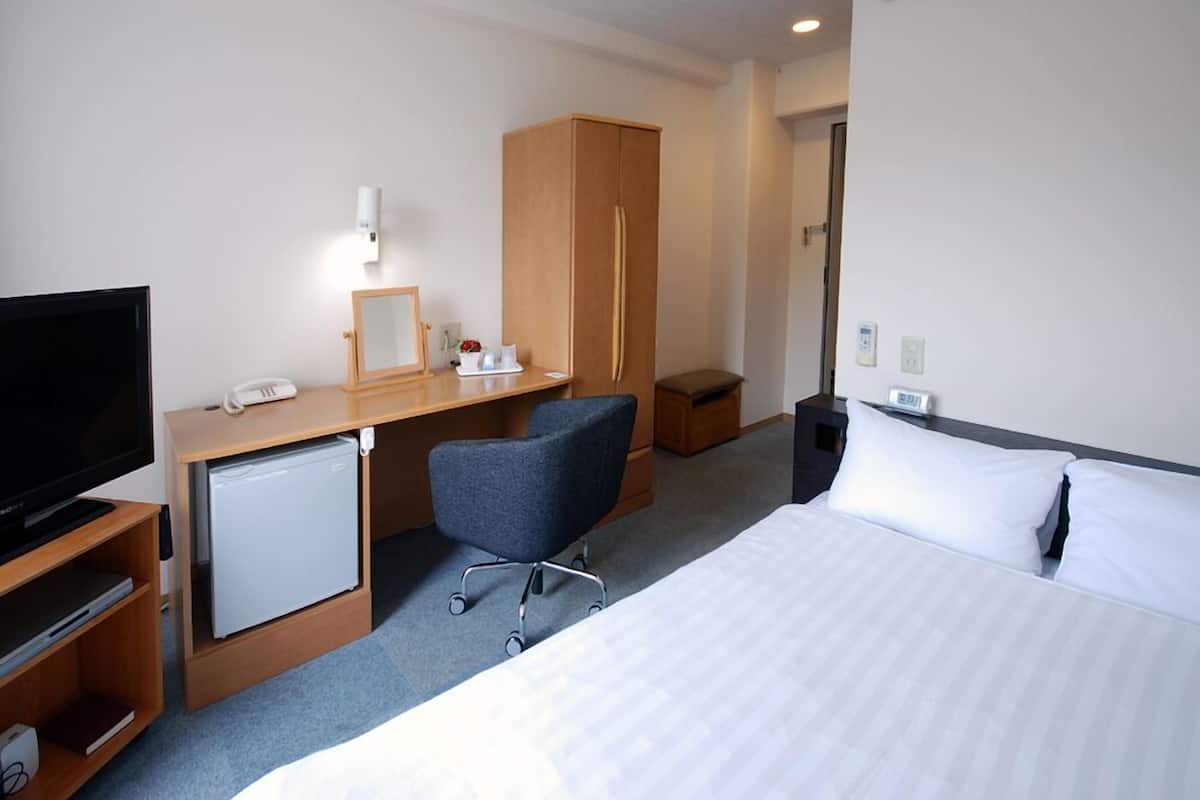 A hotel room with a bed, a desk with a computer, a television, a small refrigerator, a chair, and a wardrobe.