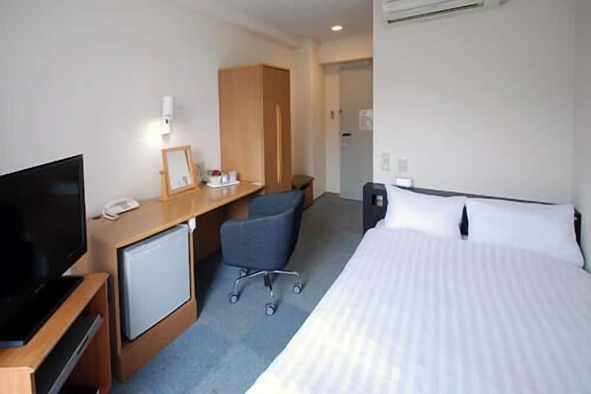 A hotel room with a bed, a desk with a computer, a chair, a TV, and a small refrigerator.
