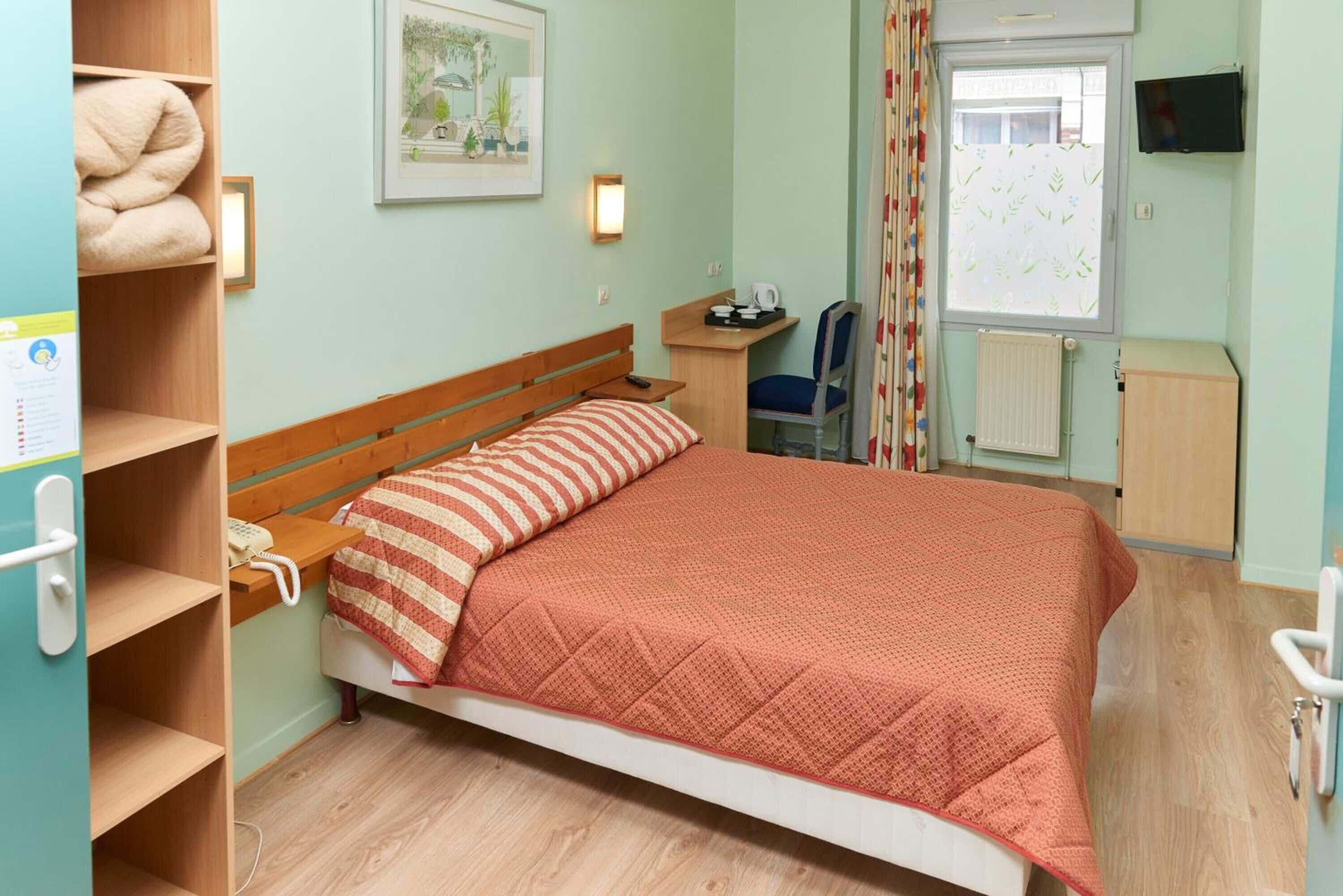 Habitación individual con una cama de una sola plaza, cabecera de madera, colcha naranja acolchada, mesita de noche con lámpara, una silla, un pequeño escritorio con silla, radiador y televisión en la pared.