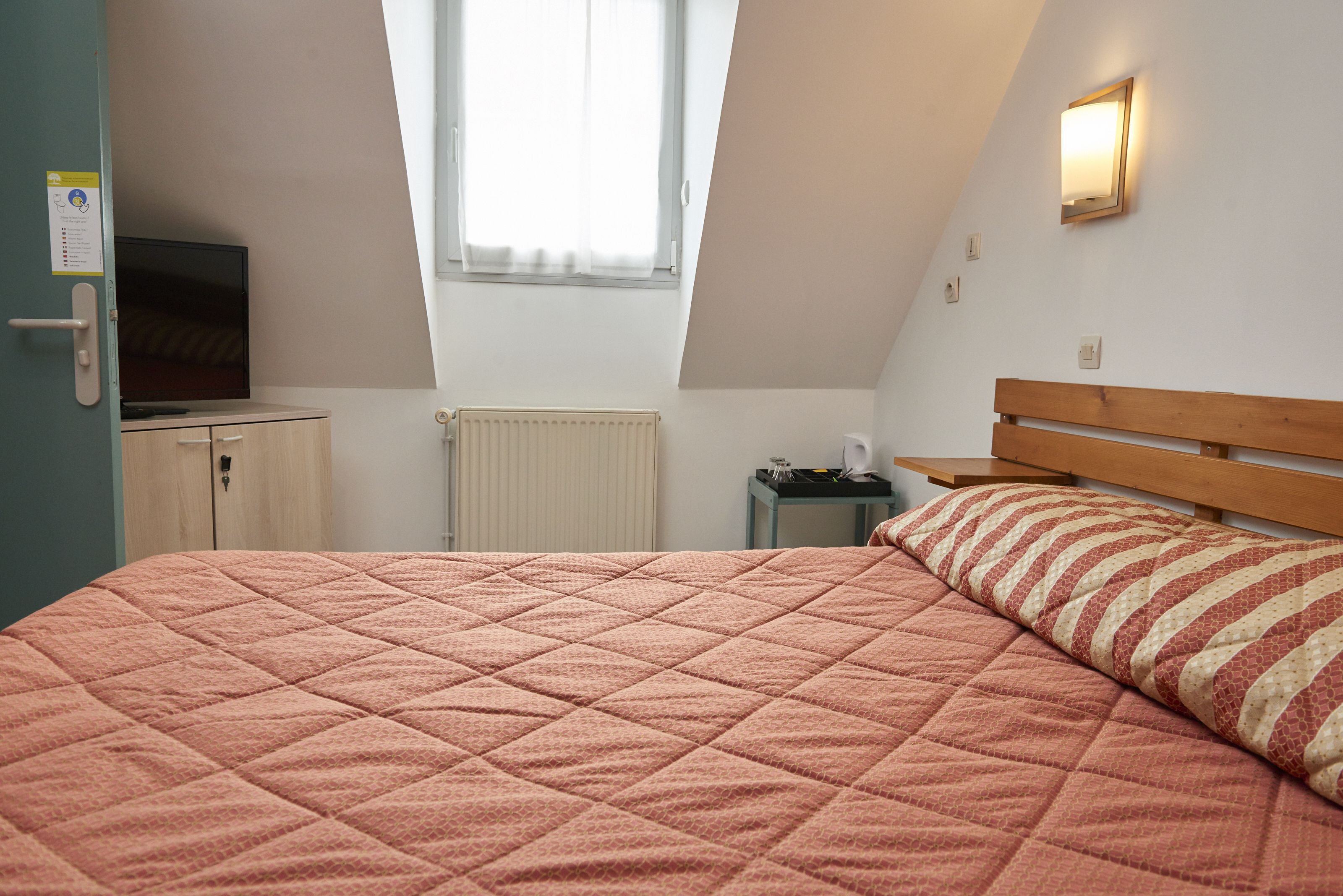 Une chambre à coucher avec un lit, une tête de lit en bois, une fenêtre, un radiateur, une petite table et un téléviseur posé sur un meuble.