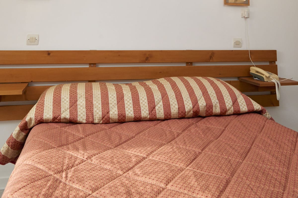A bedroom with a wooden headboard, a bed with a patterned quilt, two wall-mounted lights, and a framed picture on the wall.