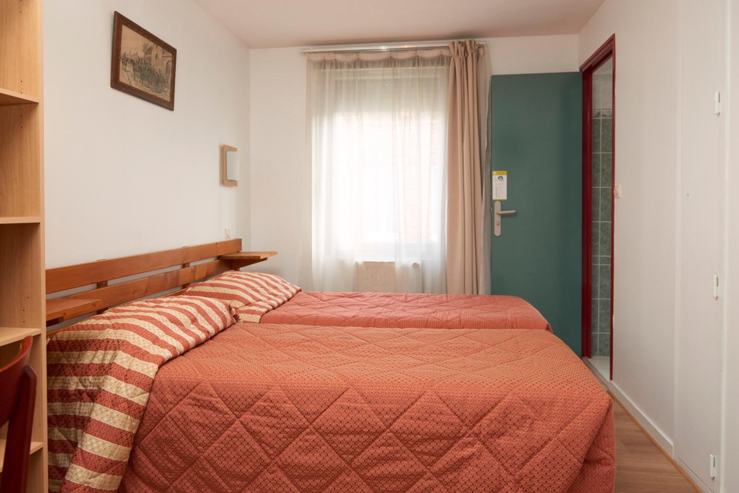 A bedroom with two beds, a wooden headboard, a window with sheer curtains, and a door with a red frame.
