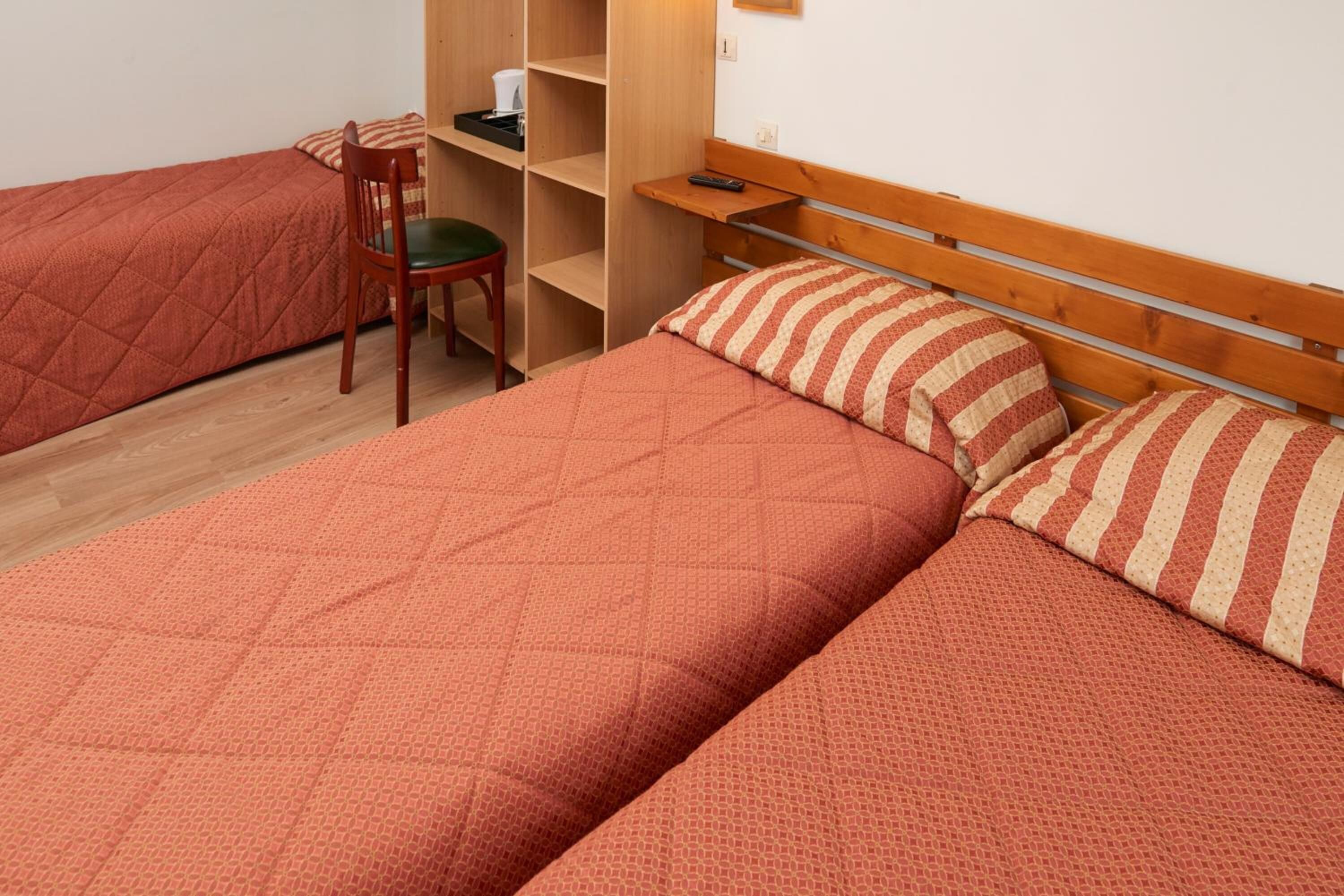 Ein Zimmer mit zwei Betten, einem Sessel, einem Regal und einem an der Wand befestigten Ablagefach.
