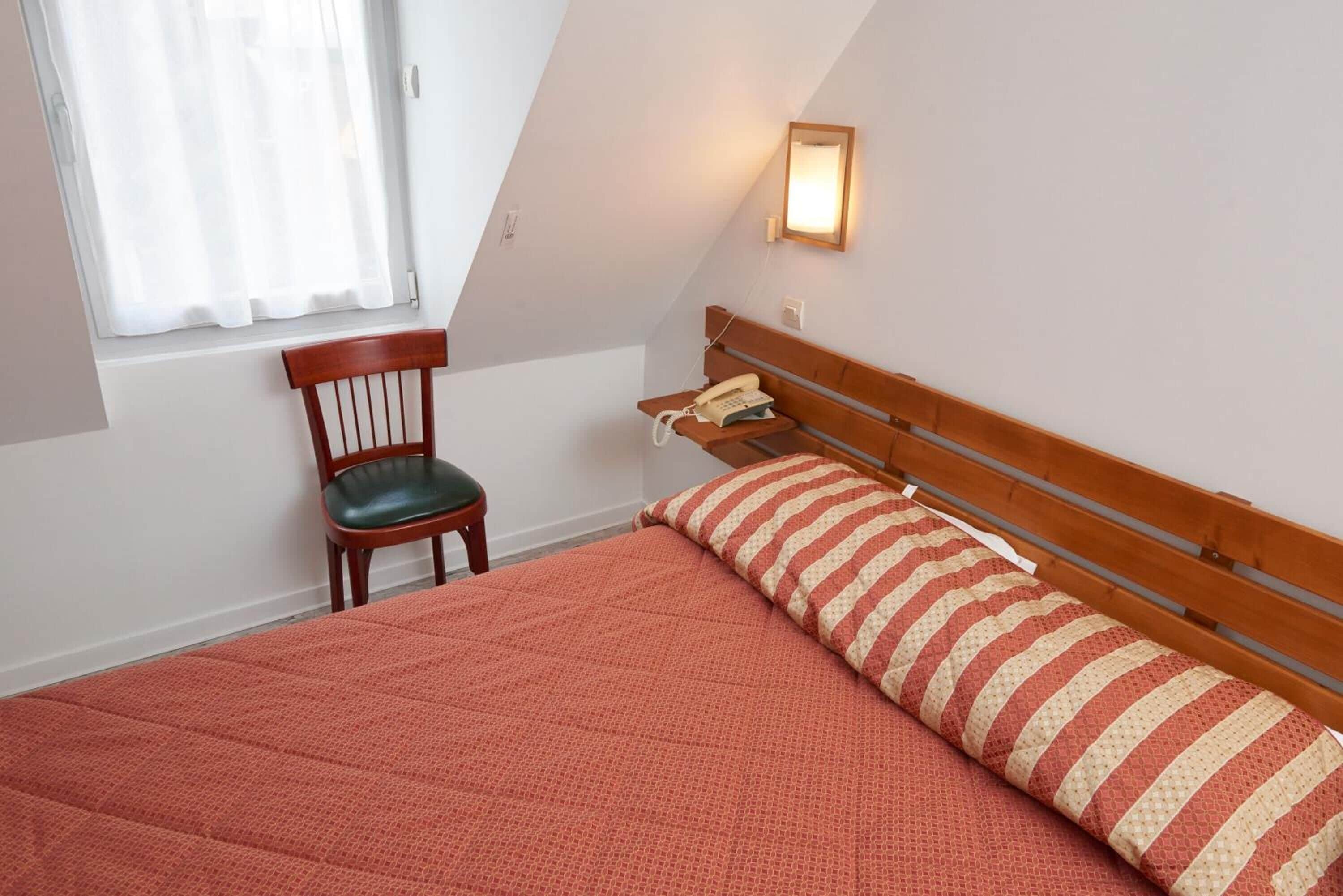 Un dormitorio con una cama, una silla, una mesita de noche con teléfono y una ventana con cortinas.