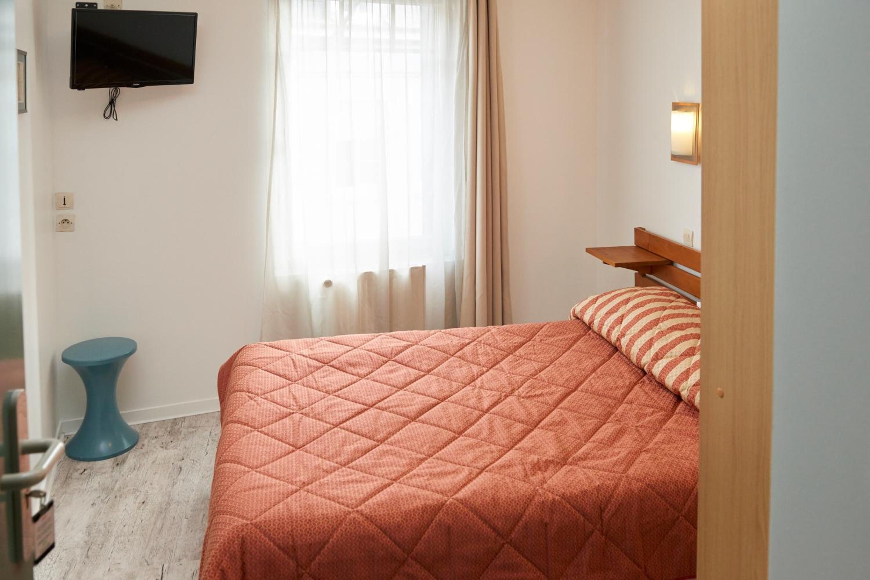 A bedroom with a bed, a TV mounted on the wall, a small blue table, and a window with curtains.