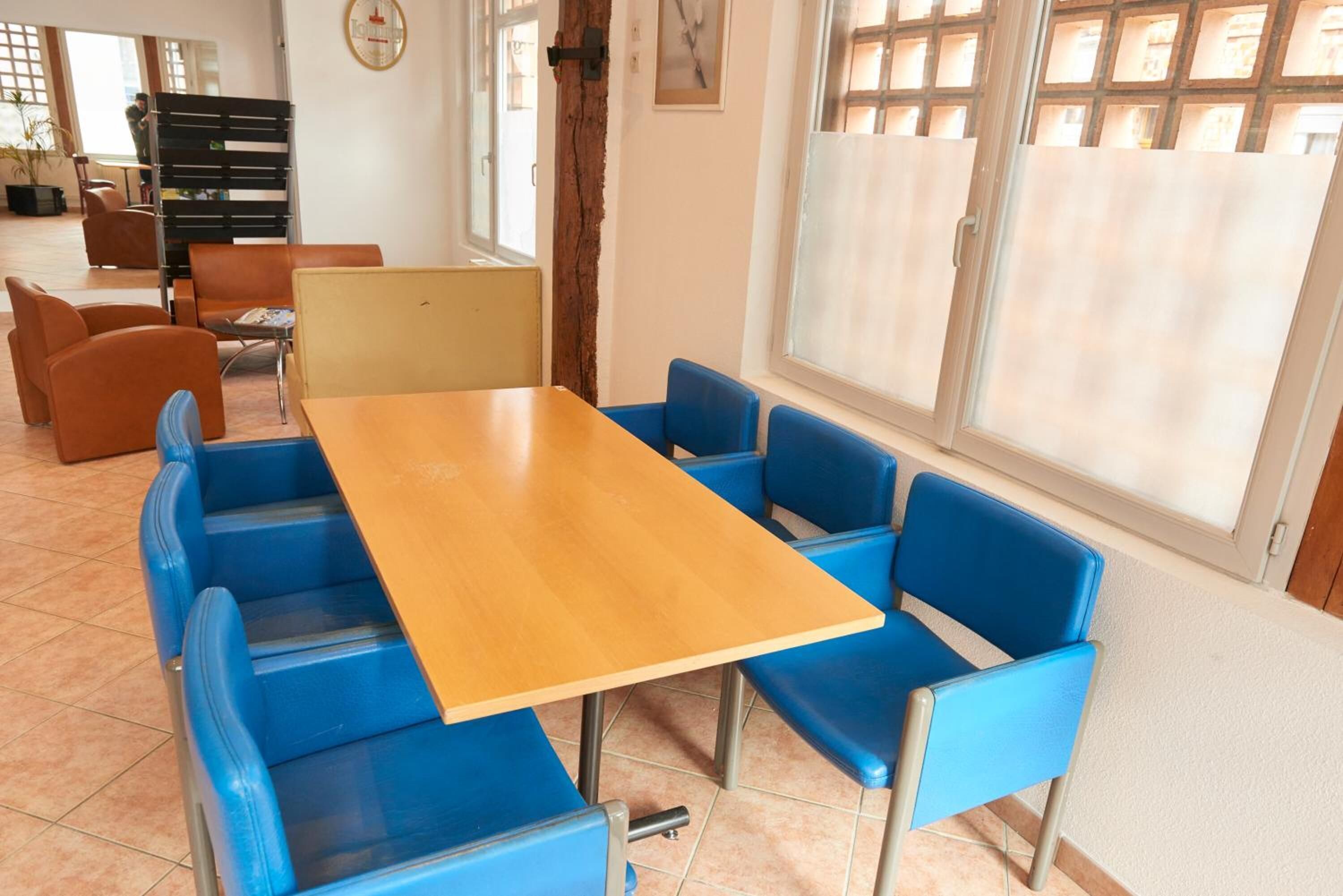 A room with a wooden table and blue chairs, a clock on the wall, and a window with blinds.