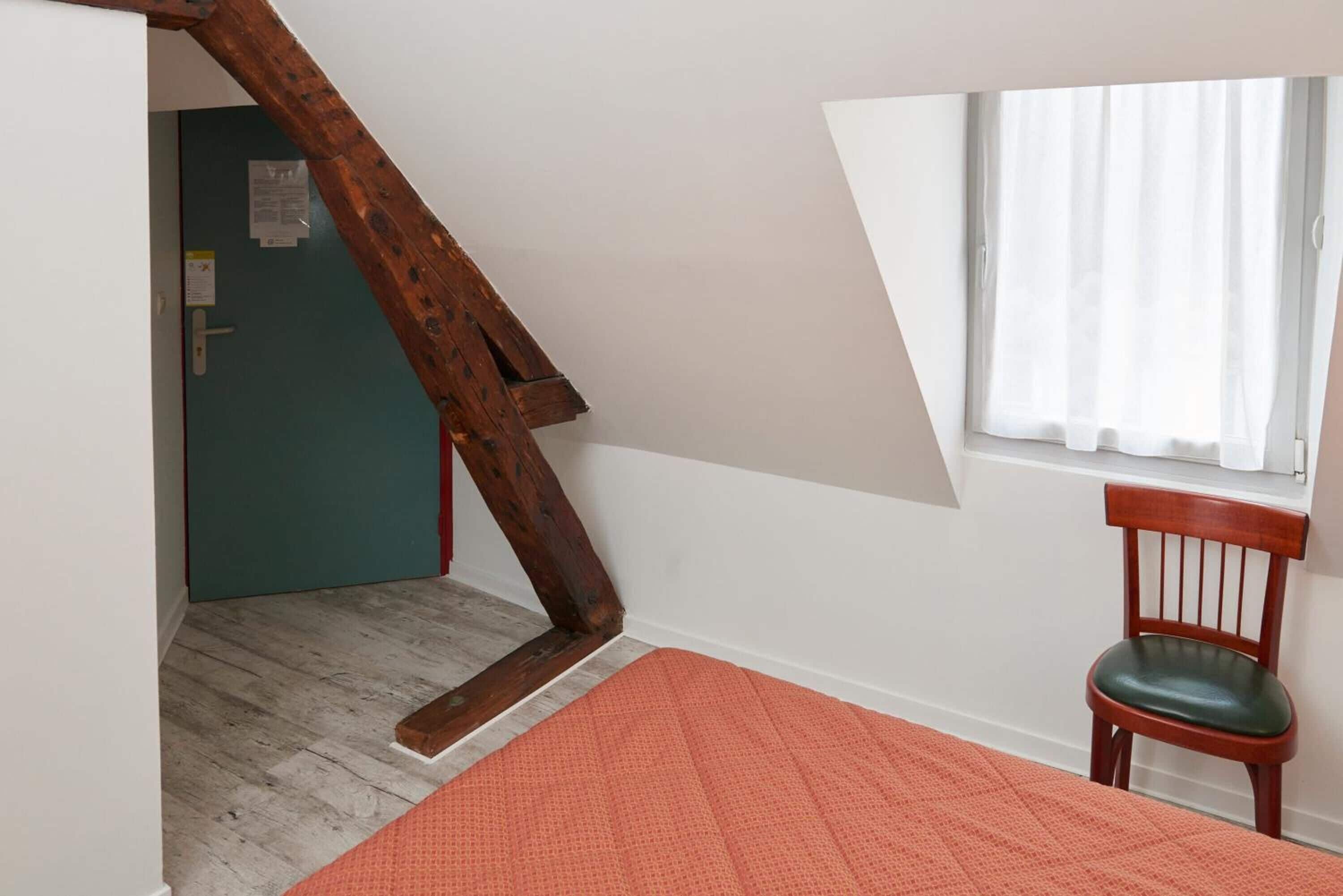 Una habitación con techo inclinado, una silla de madera, un ventanal con cortinas blancas y una puerta con marco verde.