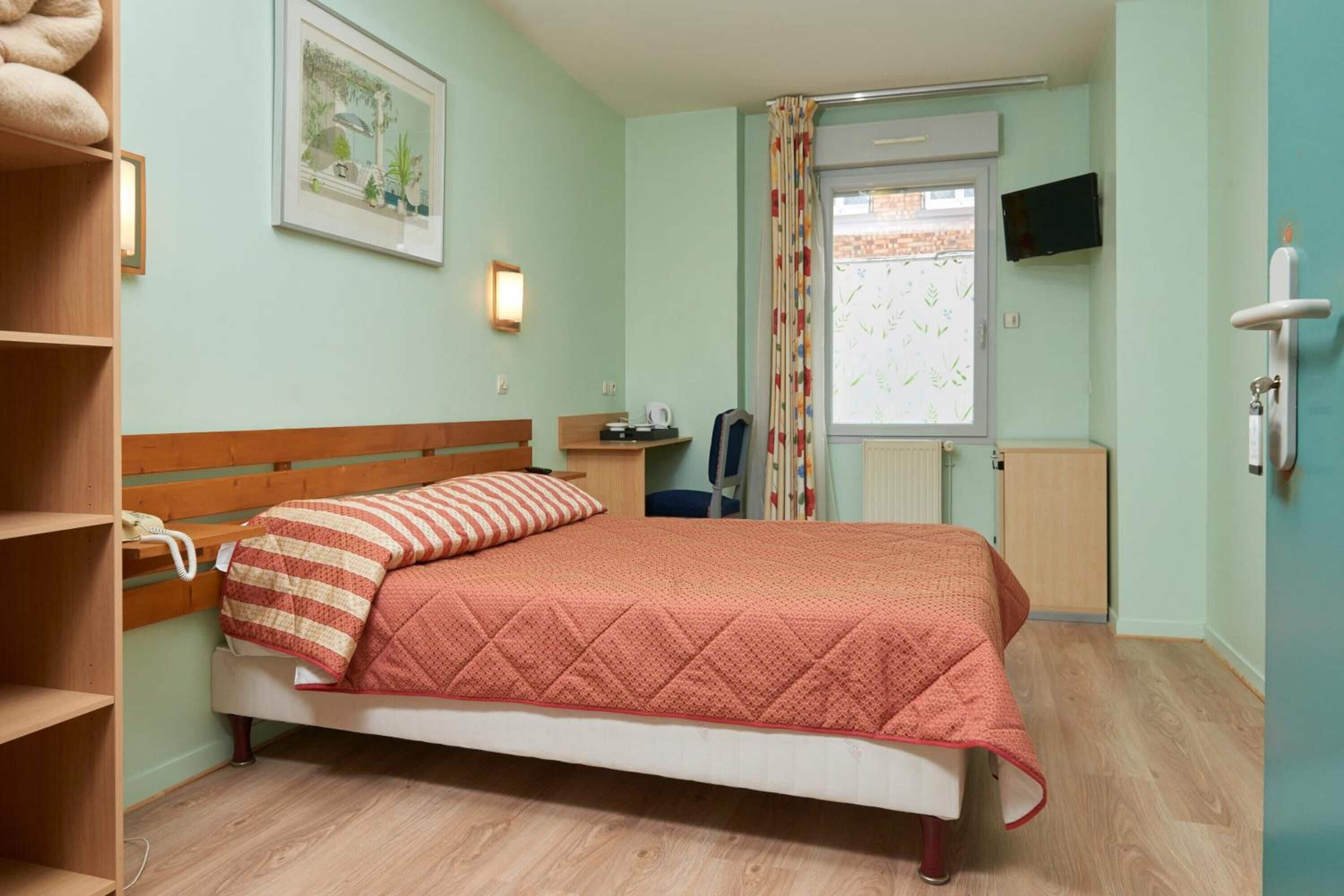 Ein Schlafzimmer mit Bett, Schreibtisch, Stuhl, Heizkörper, Fernseher und einem Fenster mit Vorhängen.