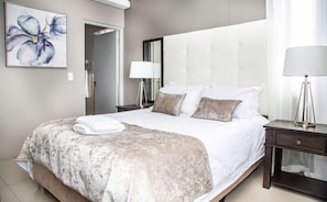 Una habitación de hotel con una cama grande, una mesita de noche con lámpara, un espejo y un cuadro de una flor en la pared.