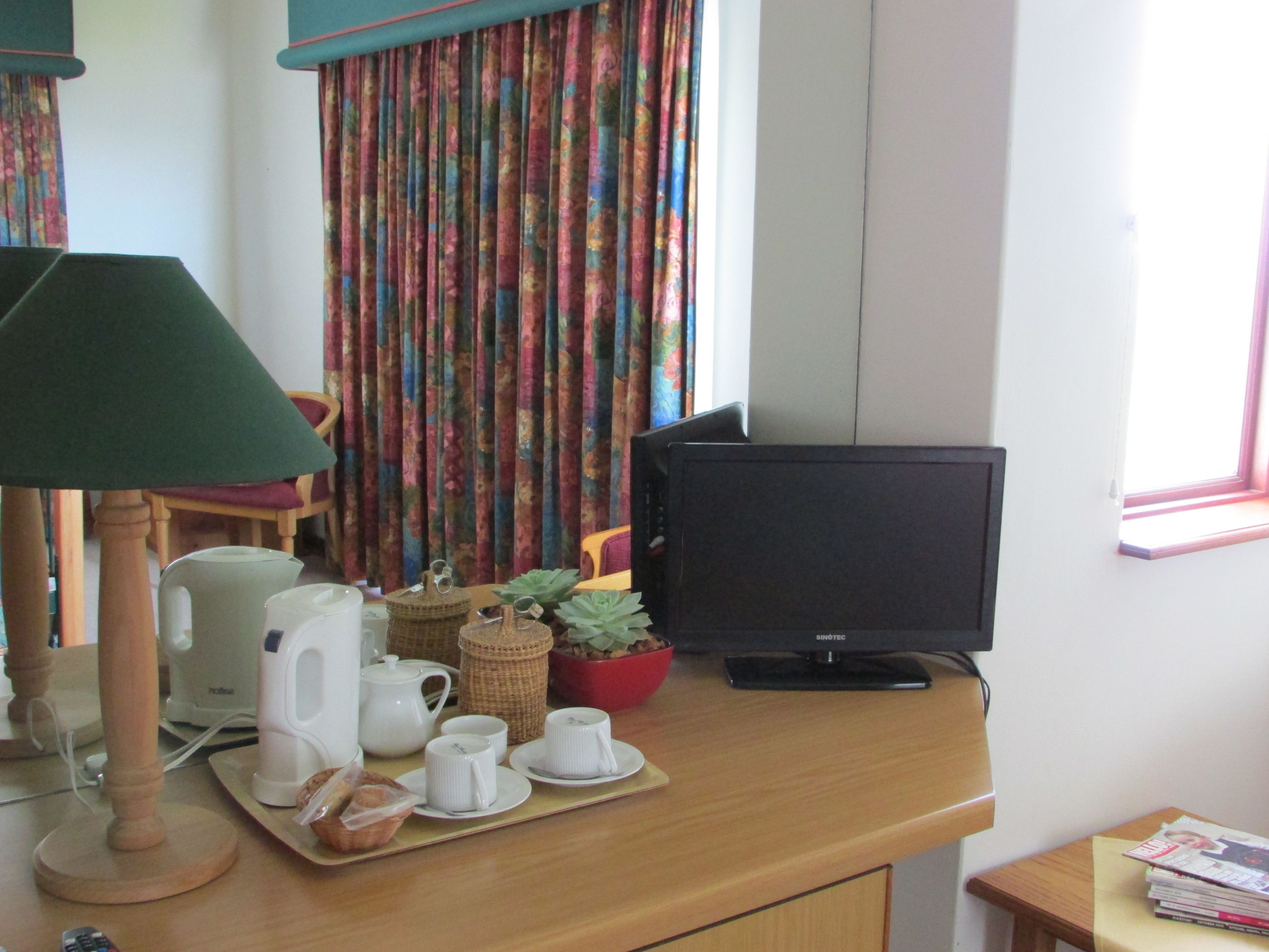 Een kamer met een bureau, een televisie, een lamp, een raam met gordijnen en een tafeltje met boeken.
