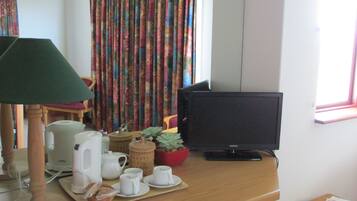 Een kamer met een bureau, een televisie, een lamp, een raam met gordijnen en een tafeltje met boeken.