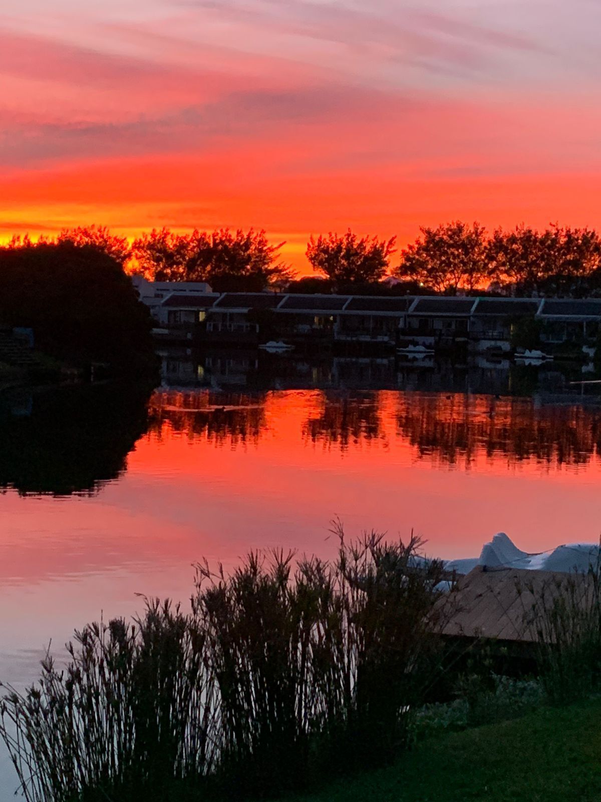 Une vue paisible sur un lac au coucher du soleil, avec une rangée de bâtiments de style traditionnel qui reflètent le ciel aux couleurs éclatantes.