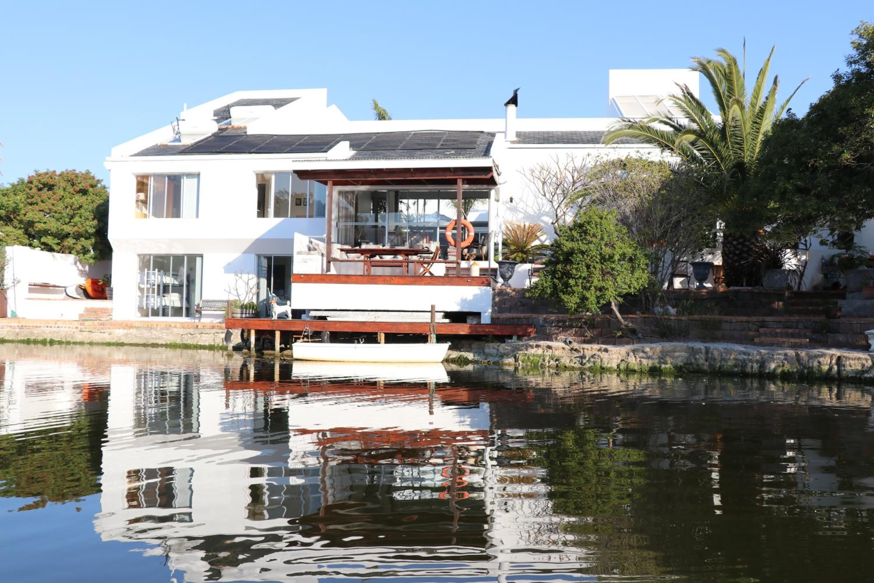 Ein modernes Haus mit einer großen Terrasse, einem vor der Tür liegenden Boot und ruhigem Gewässer, das die Struktur spiegelt.