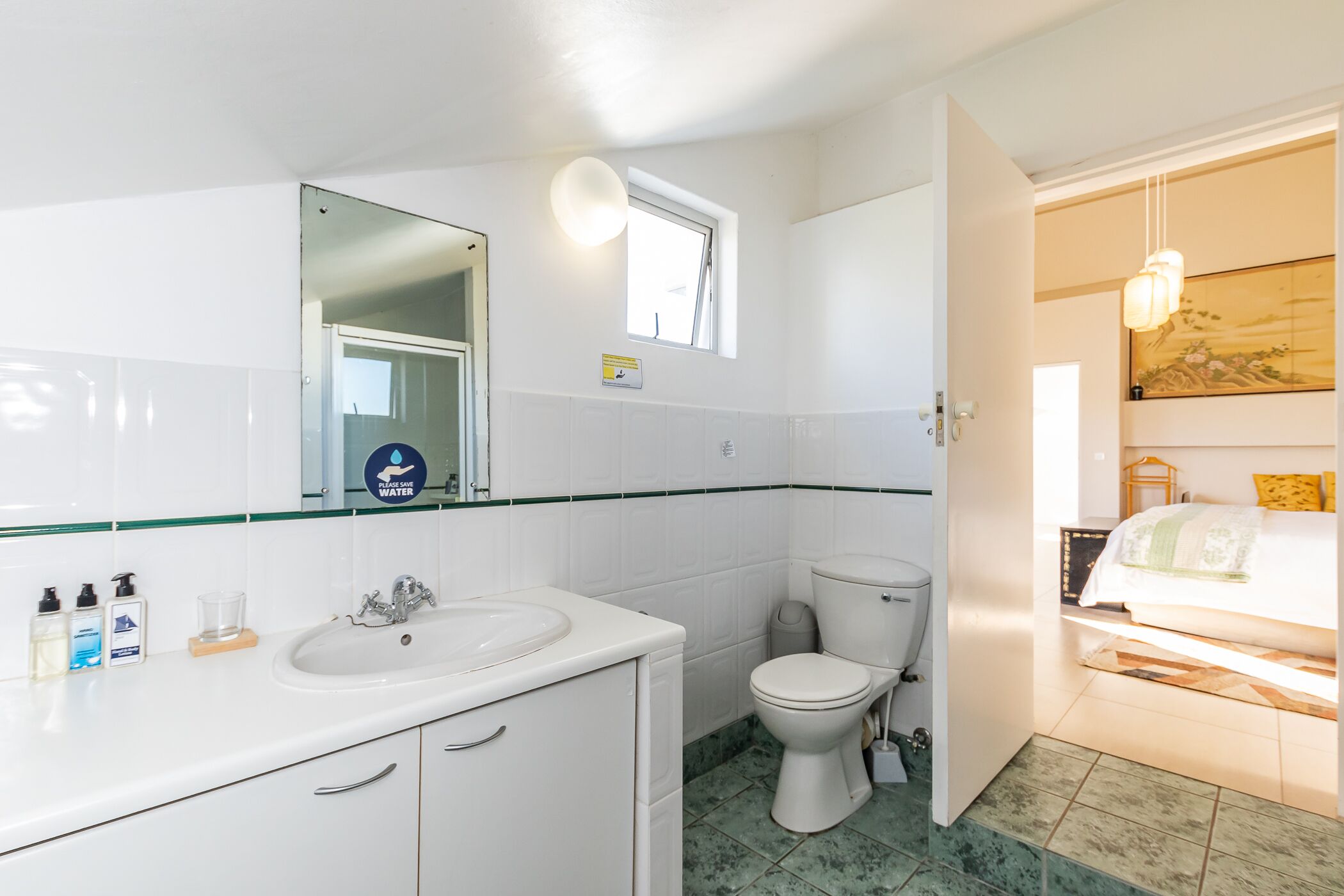 Uma casa de banho com lavatório branco, espelho e sanita. Existe uma pequena janela sobre a sanita, e vê-se uma cama no quarto adjacente.