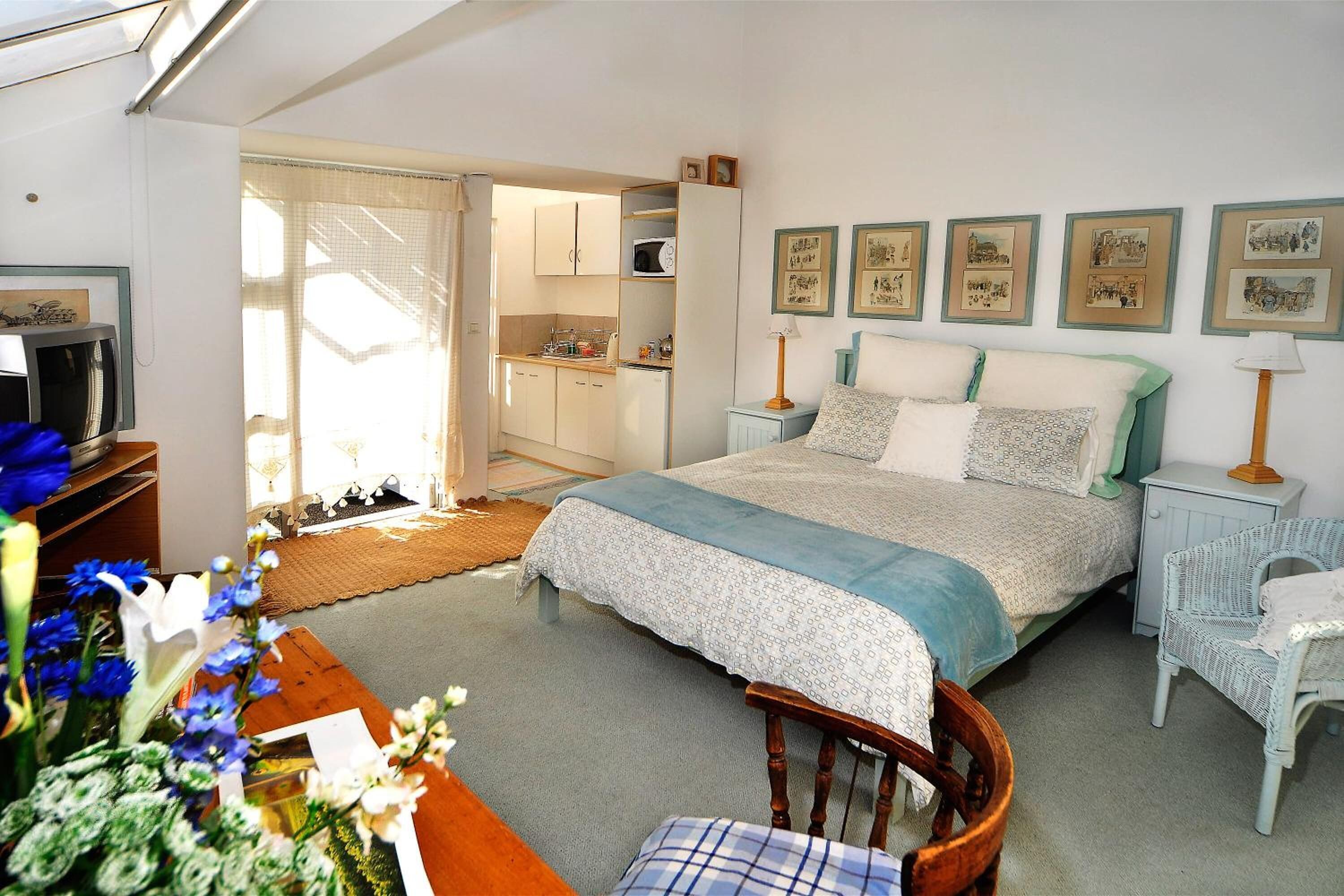 A bedroom with a bed, bedside tables, a chair, a small table, and a kitchen area visible through an open door.