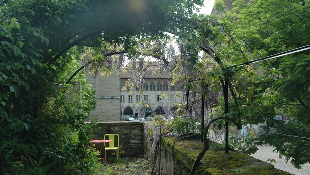 A courtyard with a stone wall, a yellow chair, and a building with arched windows in the background. - Le Clos de l'Abbaye (Cluny)