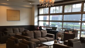 A spacious lounge area with multiple sofas and a coffee table, large windows offering a view of the outdoors, and a chandelier hanging from the ceiling. - New Station Hotel Premiere (Isahaya)