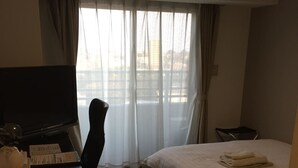 A hotel room with a bed, a desk with a computer, a chair, and a window with curtains. - New Station Hotel Premiere (Isahaya)